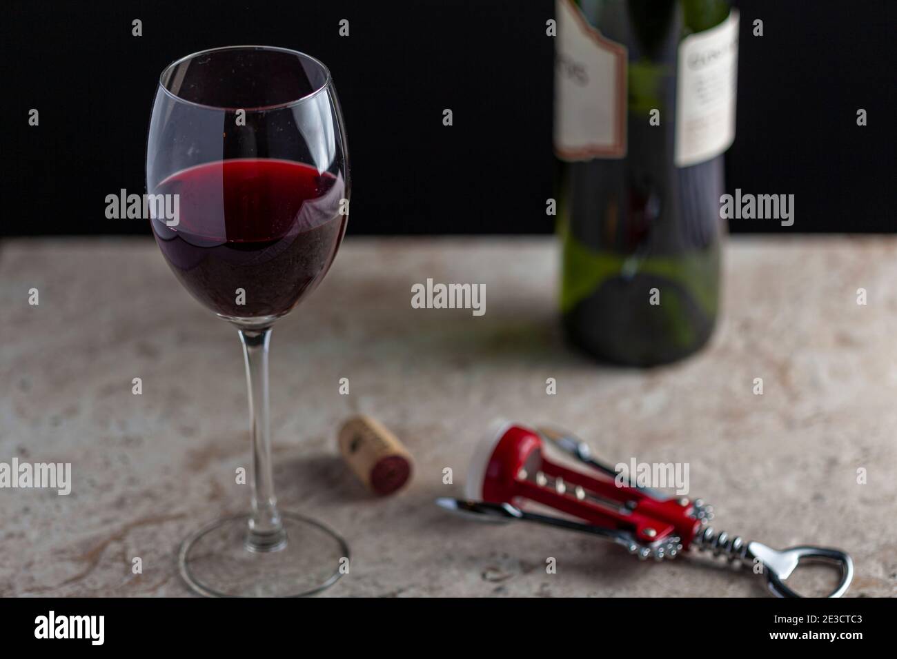 Bild eines Weinglases in Standardgröße mit Rotwein, der halbwegs gefüllt ist. Auf der Marmorplatte befindet sich ein Korkenzieher mit einem Korken daneben. Ein Gr Stockfoto