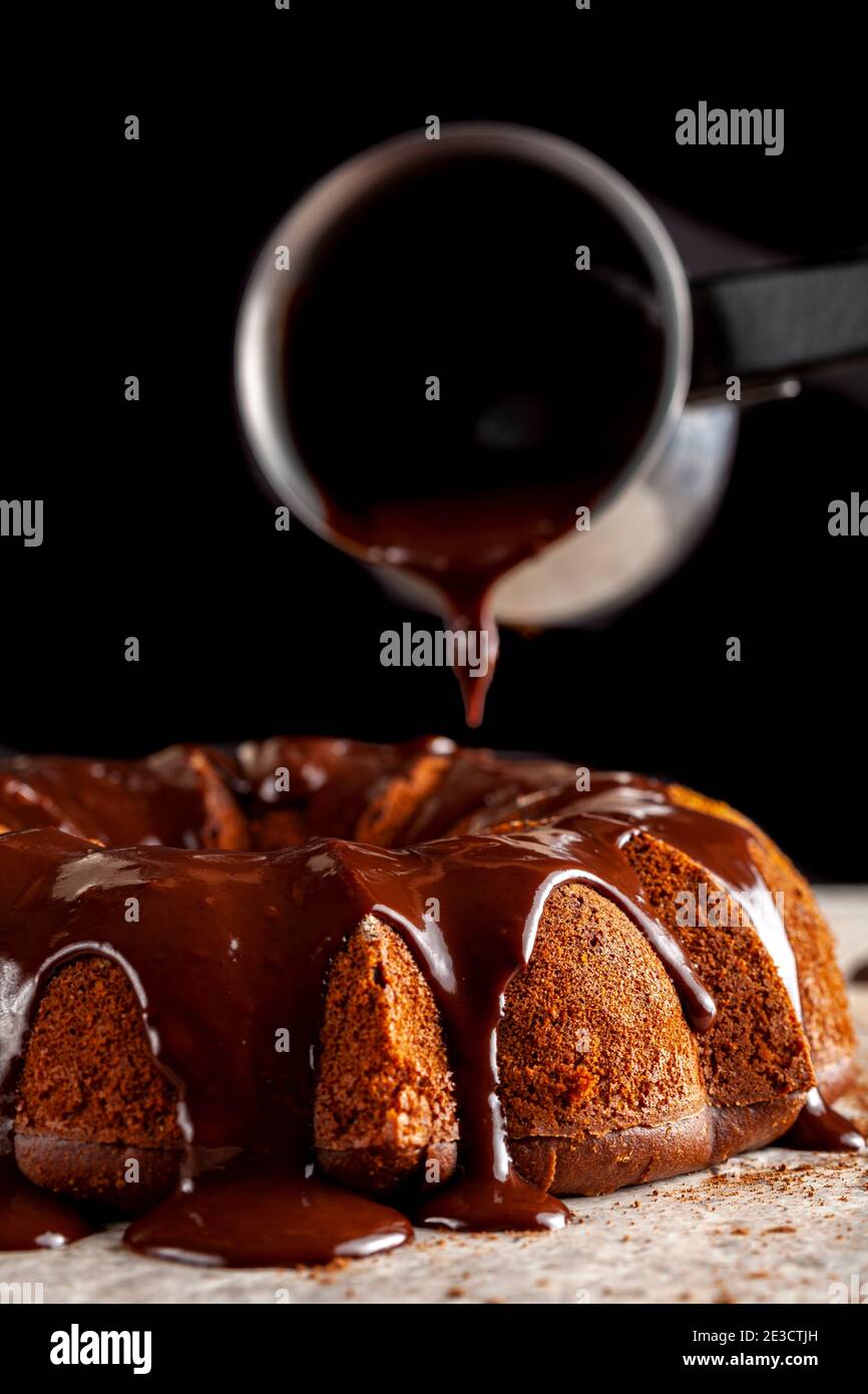 Geschmolzener heißer Schokoladenpudding wird von einem metallenen Kaffeekanne auf einen Schokoladenkuchen als Sahnehäubchen gegossen. Es fließt nach unten und macht braune Pfütze Stockfoto