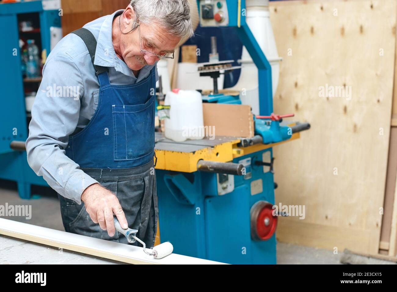 Ein älterer Tischler in Overalls und Gläsern malt ein Holzbrett mit einer Walze auf eine Werkbank in einer Schreinerei. Stockfoto