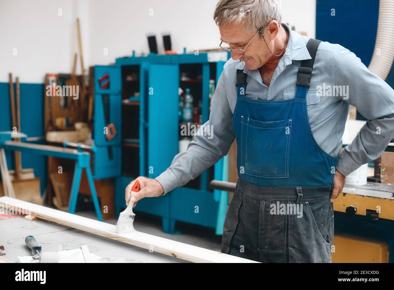 Ein älterer Tischler in Overalls und Gläsern malt in einer Schreinerei mit einem Pinsel ein Holzbrett auf eine Werkbank. Stockfoto