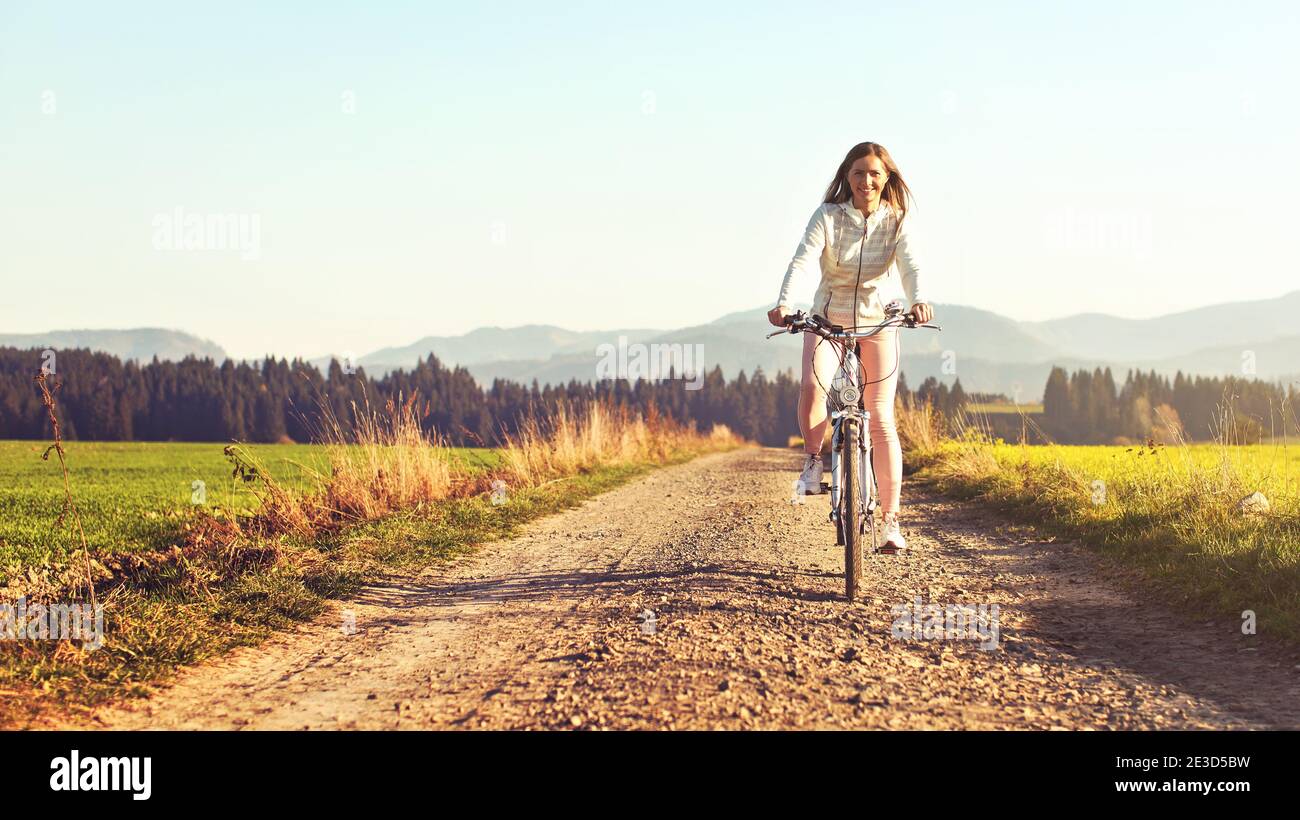 Junge Frau fährt Fahrrad auf staubiger Landstraße, Nachmittagssonne scheint auf sie, Blick von vorne Stockfoto