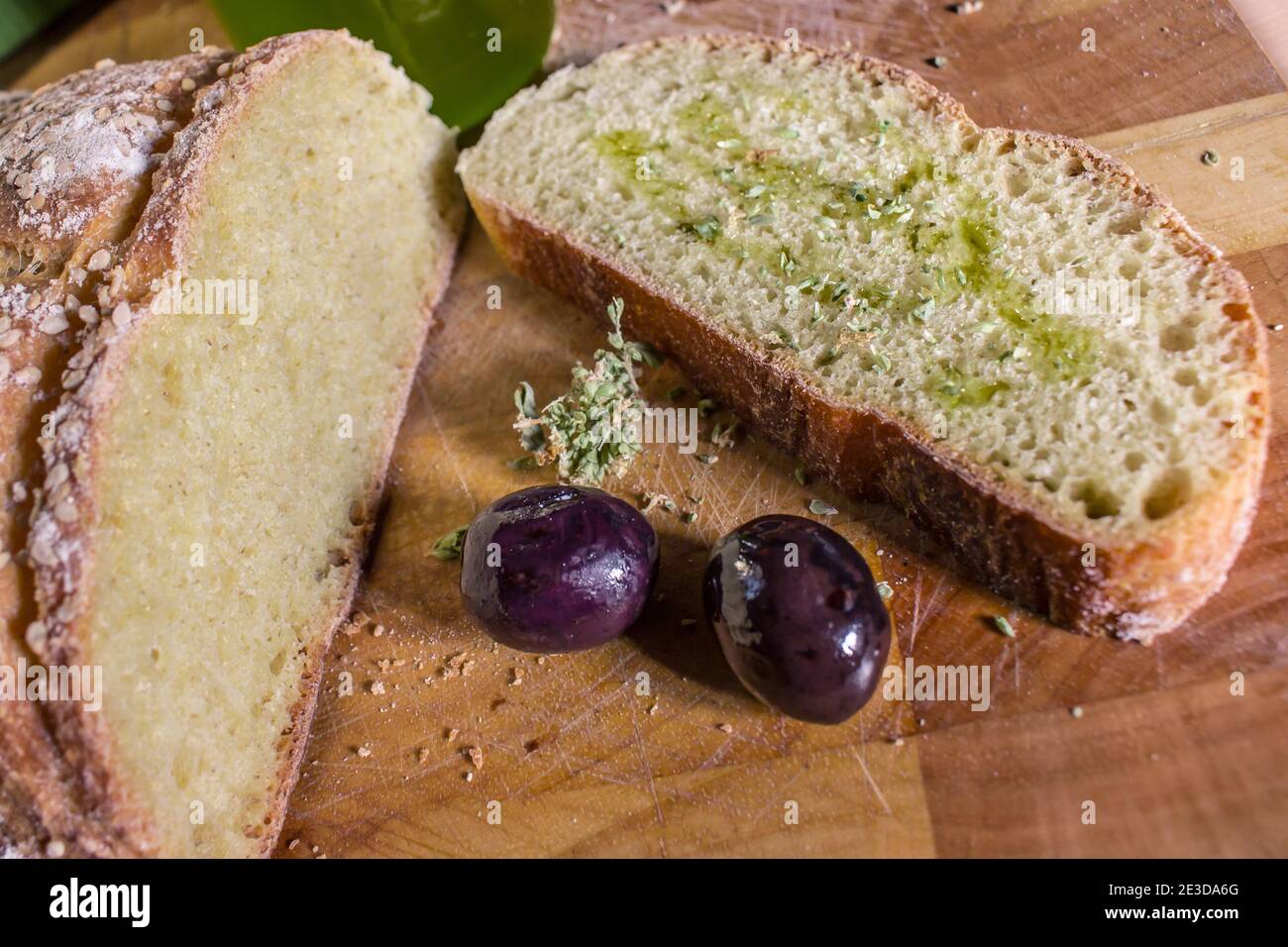 Ein Stück hausgemachtes Brot mit Olivenöl und Oregano Oben und zwei schwarze Oliven auf dem Schneidebrett Stockfoto