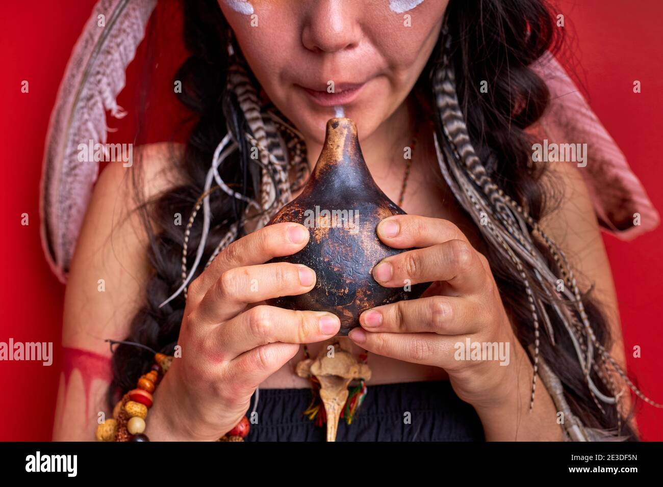 Beschnittene Schamane Frau mit authentischen indischen Pfeife isoliert auf rotem Hintergrund, weiblich mit Federn auf Kopf Schamanen. Portrait close-up Hälfte des Gesichts Stockfoto