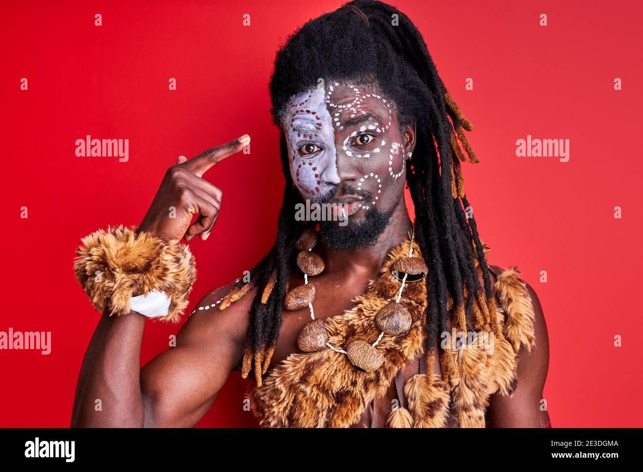 afrikanischer Mann in ethnischer Abnutzung, der Finger am Kopf färbt, steht in Schock, redet etwas, isoliert über rotem Hintergrund Stockfoto