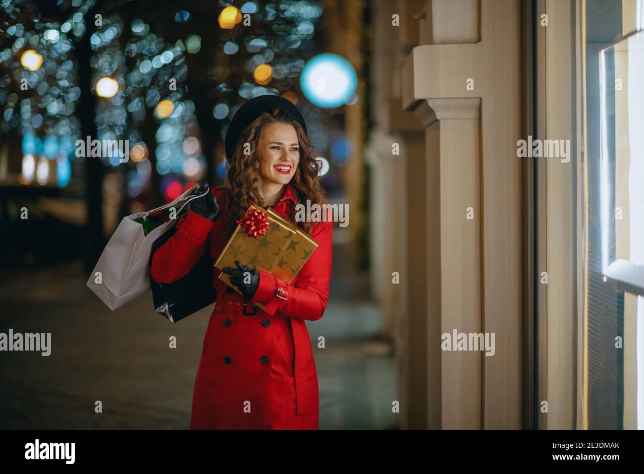 Winterspaß. Glückliche moderne Frau Shopper in roten Mantel und schwarze Baskenmütze mit Einkaufstaschen und Geschenke Blick auf das Schaufenster draußen in der Stadt in der Stockfoto
