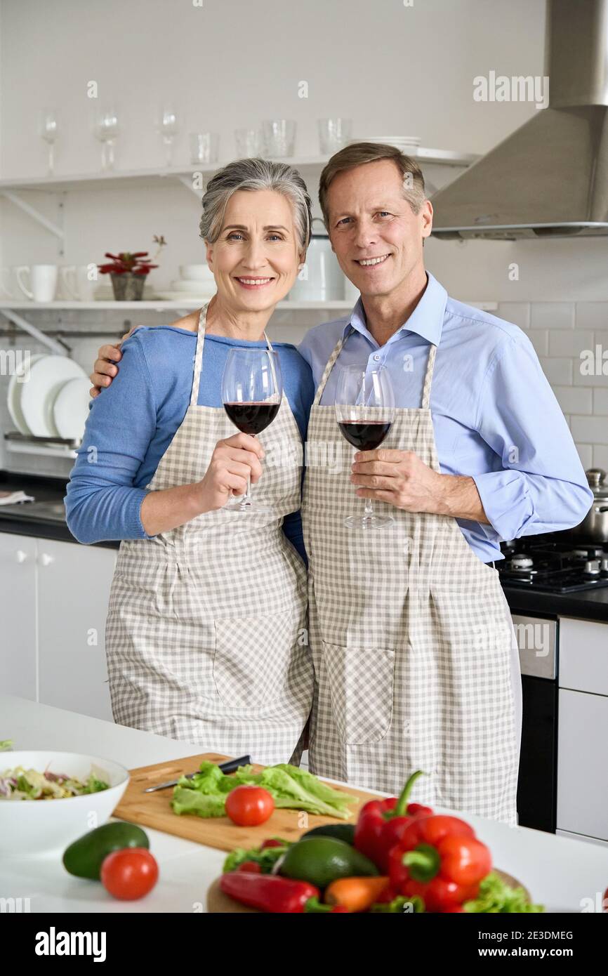 Glückliches altes Paar in Schürzen umarmend, Wein trinkend, Blick auf Kamera in der Küche. Stockfoto