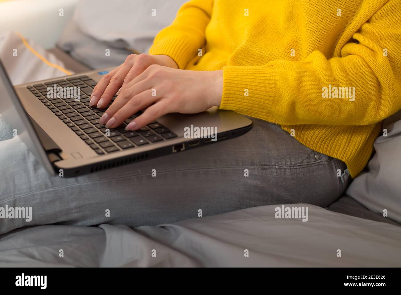 Weibliche Hände tippt Text auf einem Laptop, der auf dem Bett sitzt. Arbeiten Sie von zu Hause Trendfarben von 2021 - Beleuchtung gelb ultimative graue Farbe Stockfoto