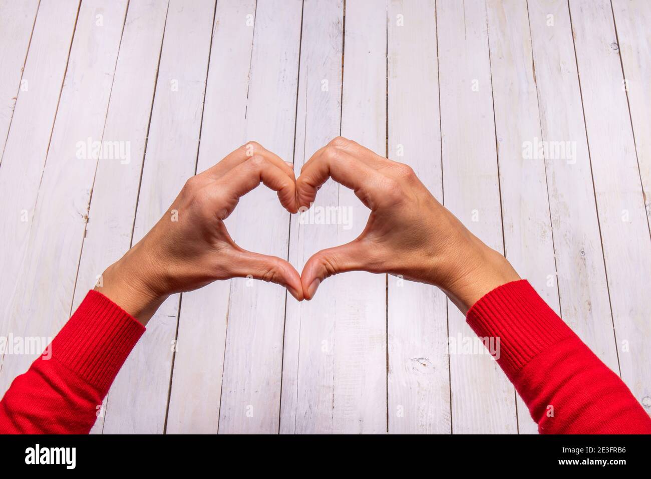 Weibliche Hände machen eine Herzform auf einem weißen Holzhintergrund. Ein Hauch von Rot aus dem Pullover zieht die Aufmerksamkeit auf sich. Leeres Leerzeichen für Valentine-Text. Stockfoto