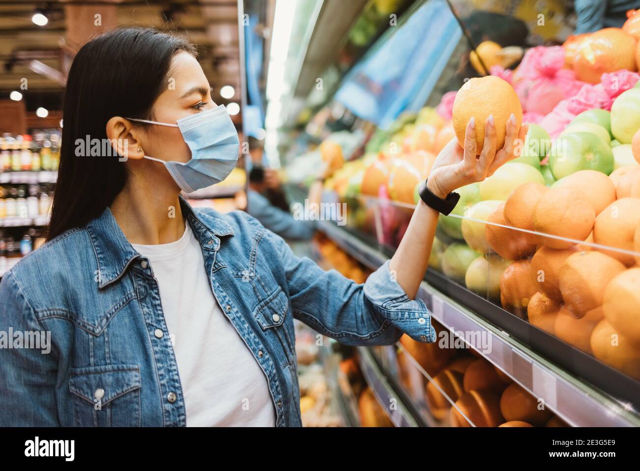 Junge Frau in Schutzmaske und Jeansjacke kauft Obst Im Geschäft Stockfoto