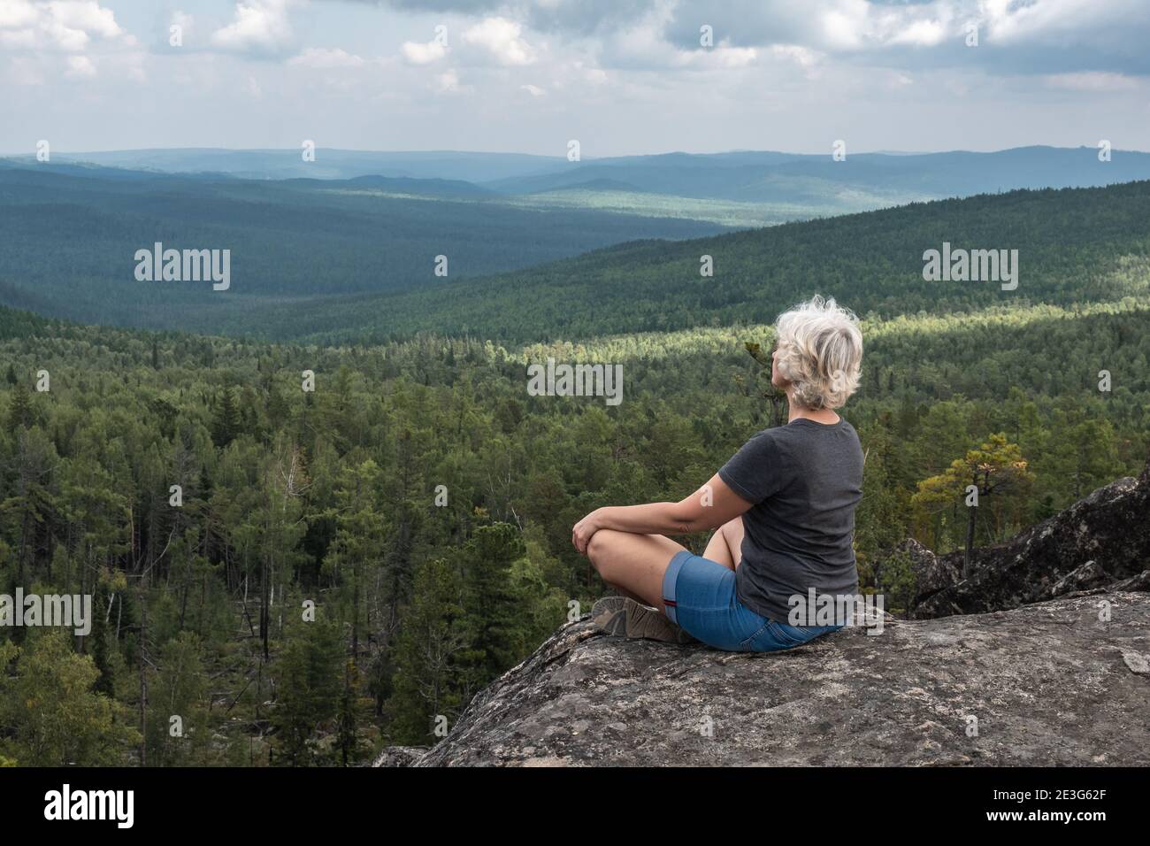 Eine Frau mittleren Alters praktiziert Yoga und Meditation auf der Bergspitze mit herrlichem Blick auf bewaldete, hügelige Tal. Ruhe und Entspannung. Gesunder Lebensstil Stockfoto