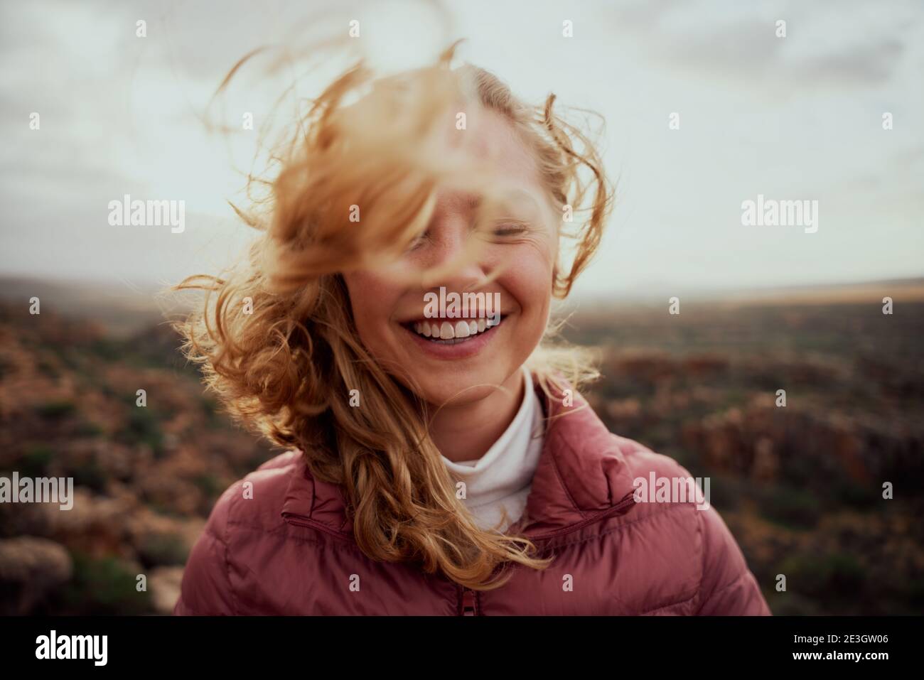 Porträt der jungen lächelnden Frau Gesicht teilweise mit Fliegen bedeckt Haare in windigen Tag stehen am Berg - unbeschwerte Frau Stockfoto