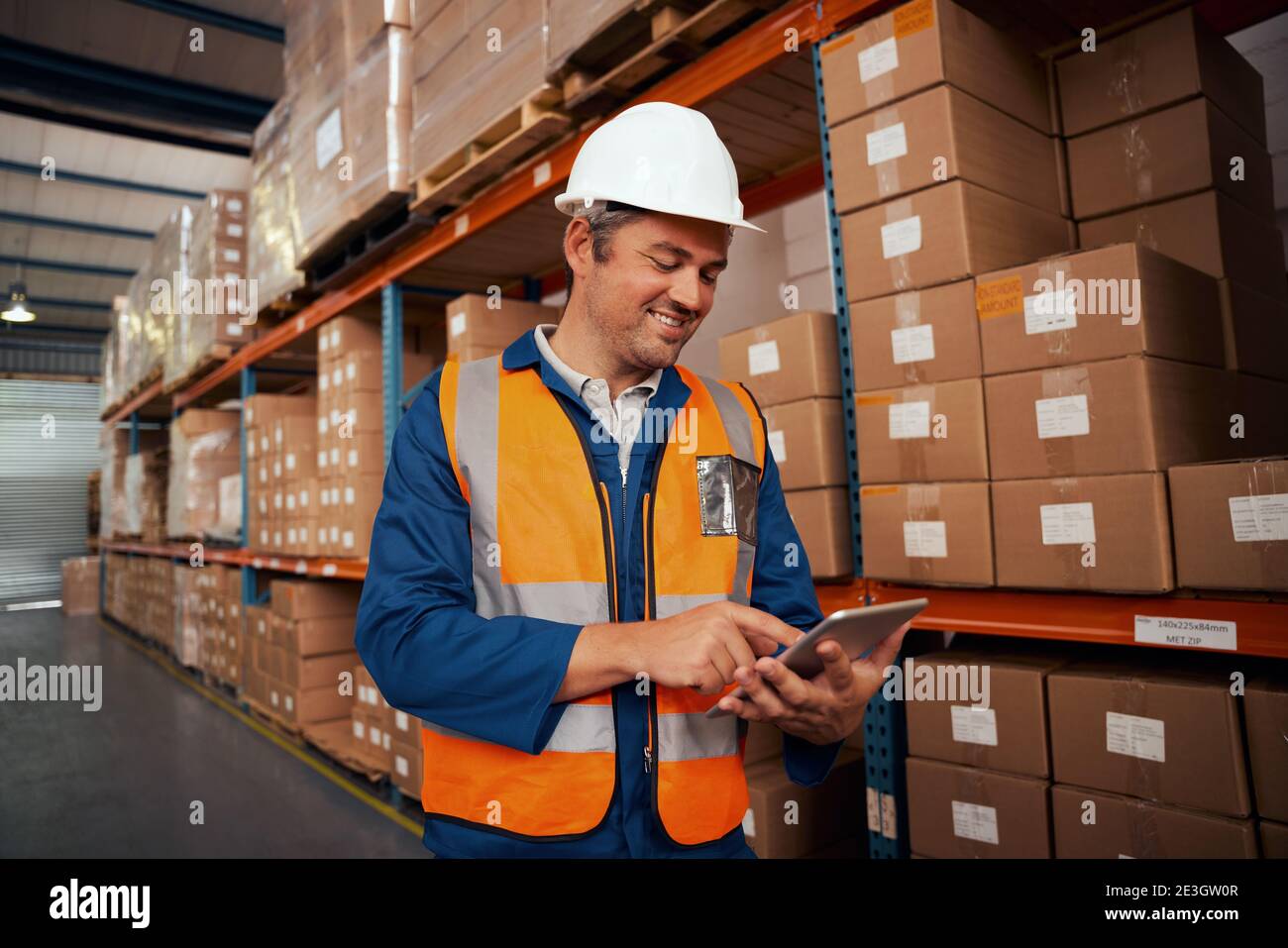 Glücklicher männlicher Arbeiter, der in der Fabrik mit einem digitalen Tablet im Stehen arbeitet In der Nähe des Stapels von Kartons Stockfoto