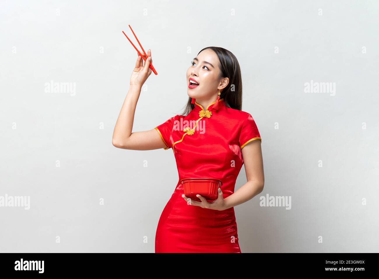 Asiatische Frau in traditionellen roten chinesischen Qipao Kleid hält Kunststoff Lebensmittelkarton und Essstäbchen, die in isoliertem Licht nach oben schauen Grauer Hintergrund Stockfoto