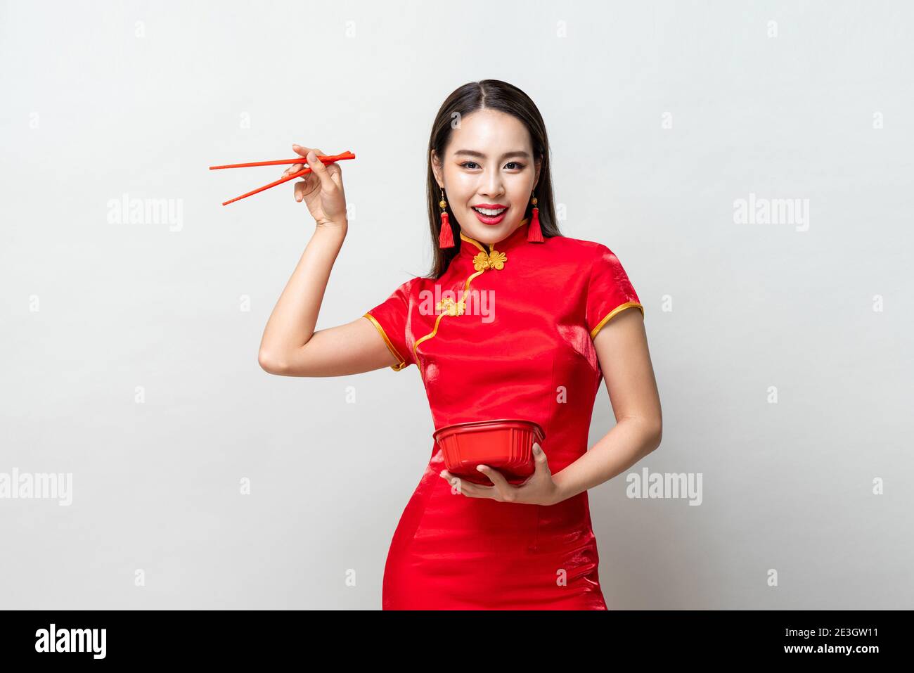 Hübsche asiatische Frau in traditionellen roten chinesischen Qipao Kleid halten Kunststoff-Lebensmittelbox und Essstäbchen in Studio isoliert grauen Hintergrund Stockfoto