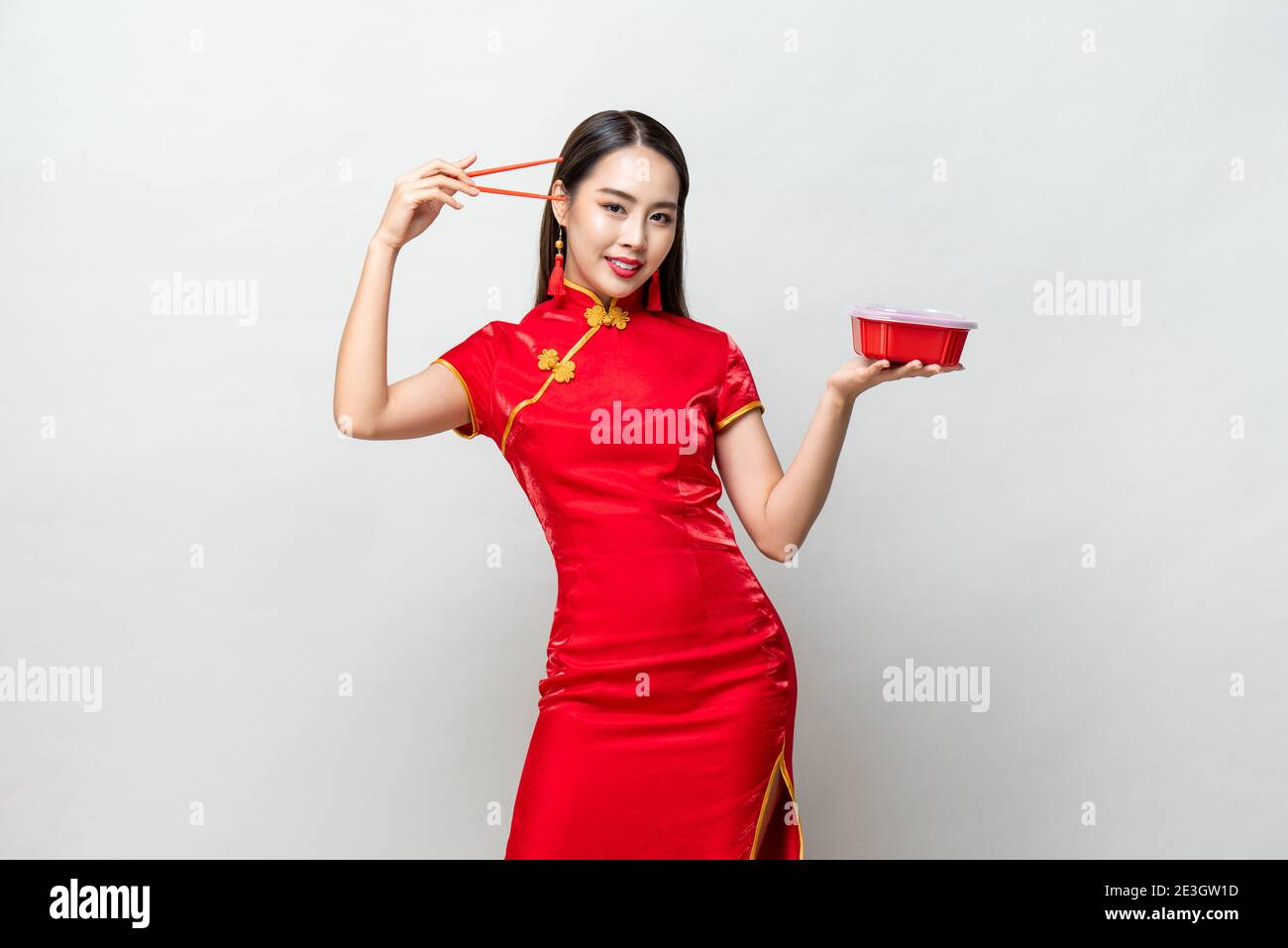Hübsche asiatische Frau in traditionellen roten chinesischen Qipao Kleid halten Kunststoff-Lebensmittelbox und Essstäbchen in Studio isoliert grauen Hintergrund Stockfoto