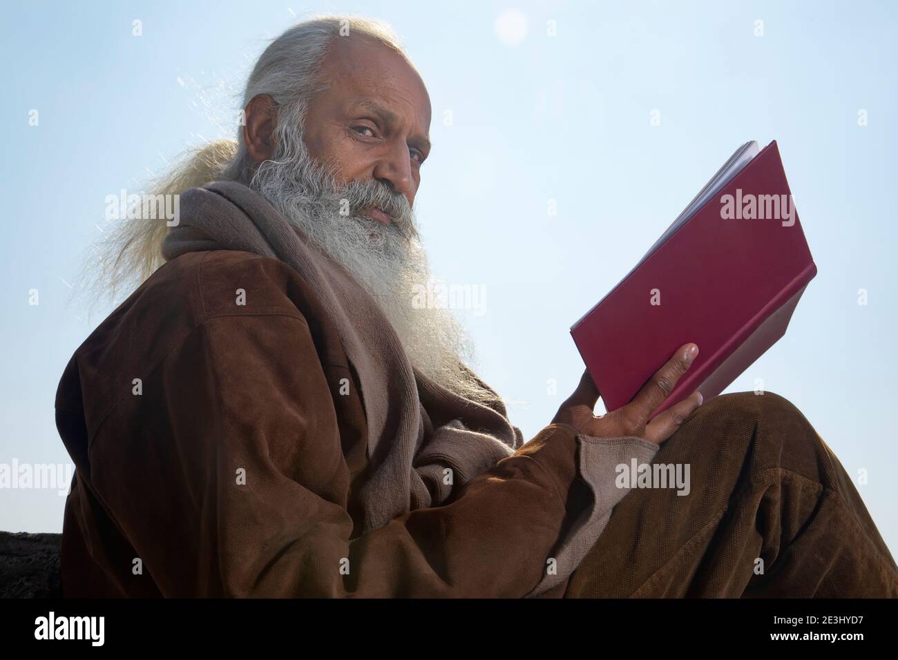 EIN BÄRTIGER ALTER MANN, DER BEIM LESEN DES BUCHES DIE KAMERA ANSAH Stockfoto