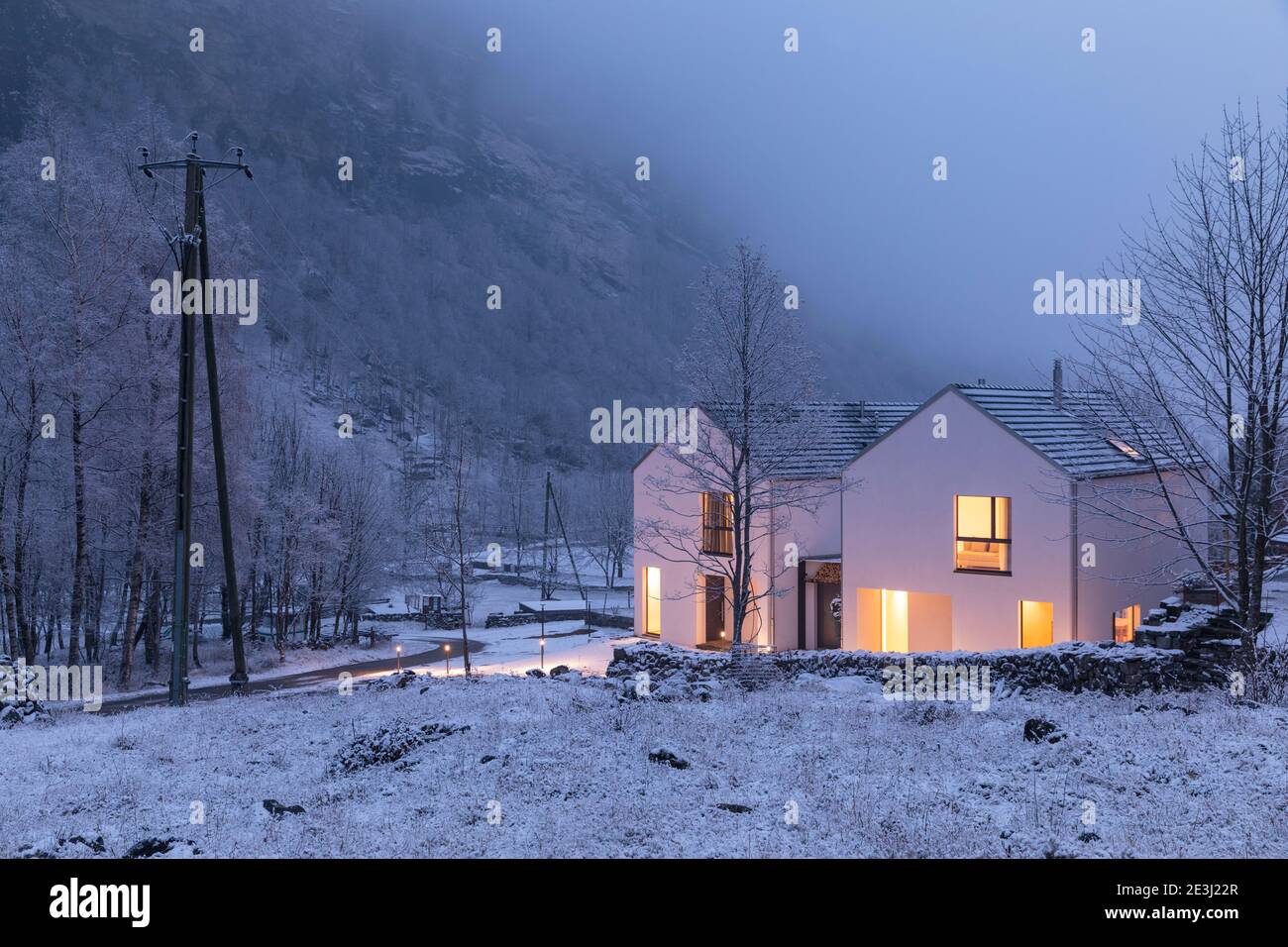 Modernes weißes Haus mit hellen Fenstern umgeben von Bergen, Schnee und Bäumen. Chirstmas Stimmung. Niemand drinnen. Stockfoto
