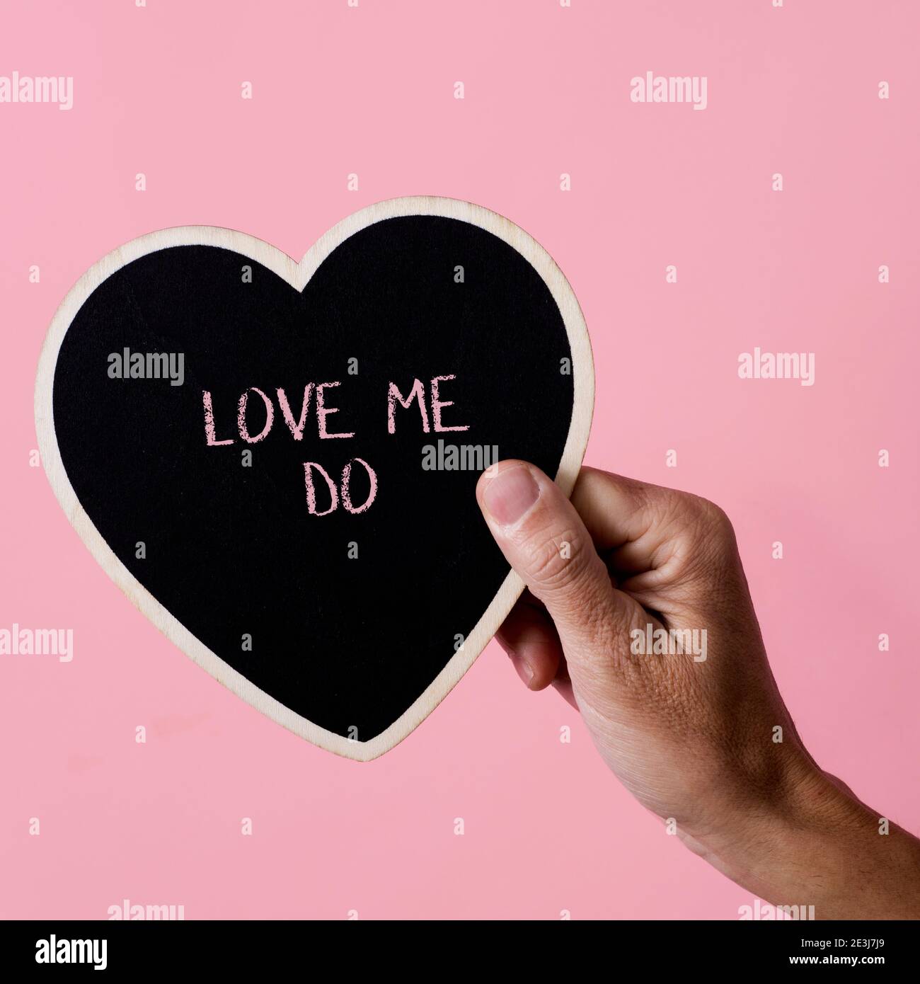 Nahaufnahme eines Mannes, der ein schwarzes herzförmiges Zeichen in der Hand hält, mit dem Text Love Me Do, der darin geschrieben ist, auf einem rosa Hintergrund, mit einem quadratischen Format Stockfoto