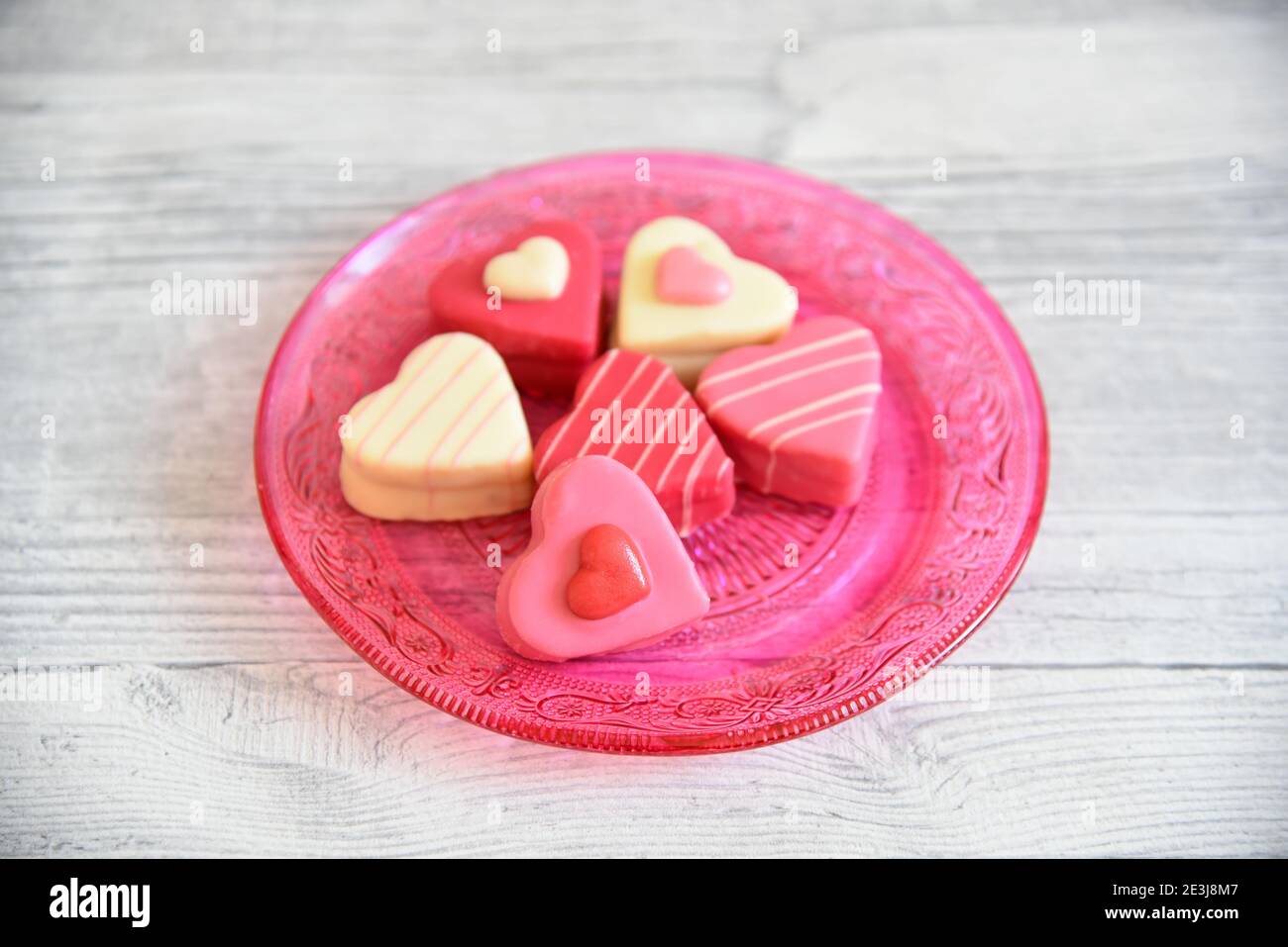 Herzförmige bunte Pralinen (Petit Fours) auf einer rosa Glasplatte mit Ornamentmuster. Heller Holztisch Hintergrund. Stockfoto