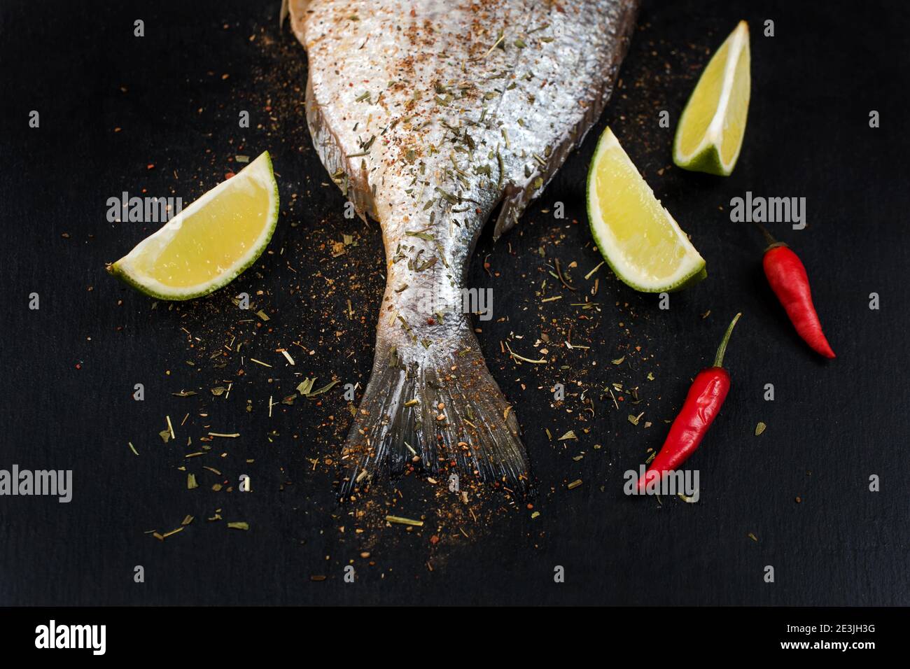 Schwanz aus frischem rohem dorado auf schwarzem Schiefersteinbrett mit Gewürzen, Kräutern, Zitrone und rotem Paprika. Brassen Fisch bereit zum Kochen. Draufsicht. Stockfoto