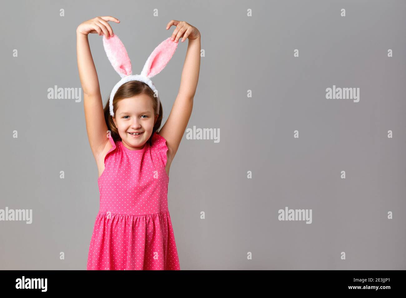 Frohe Ostern. Fröhliche kleine Mädchen in einem rosa Kleid mit Punkten auf einem grauen Hintergrund. Das Kind hält seine Hände hinter den Kaninchenohren. Stockfoto