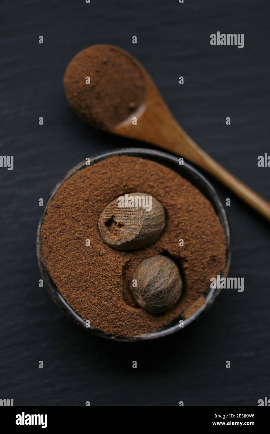 Muskatnuss.ganze und geriebene Muskatnuss in einem schwarzen Keramikbecher und Löffel auf einem schwarzen Schieferhintergrund.Gewürz- und Kräuterkonzept. Dunkle Stimmung.ganz und teilweise Stockfoto