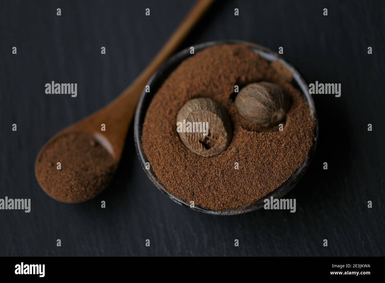 Muskatnuss.ganze und geriebene Muskatnuss in einem schwarzen Keramikbecher und Holzlöffel auf einem schwarzen Schieferhintergrund.Gewürz- und Kräuterkonzept. Dunkle Stimmung.ganz und Stockfoto