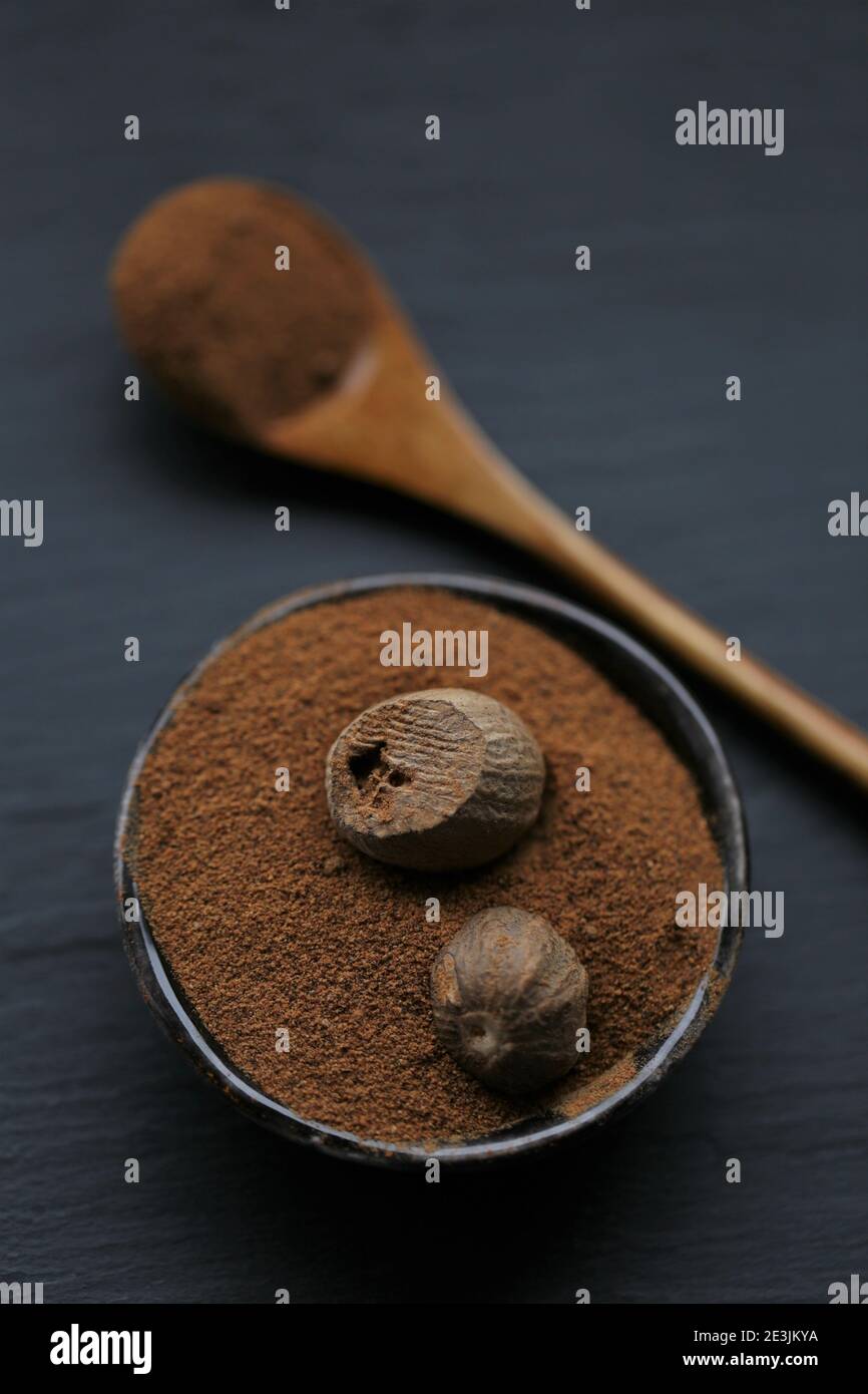 Muskatnuss.ganze und geriebene Muskatnuss in einem Keramikbecher und Holzlöffel auf einem schwarzen Schieferhintergrund.Gewürz- und Kräuterkonzept. Dunkle Stimmung.ganz und teilweise Stockfoto