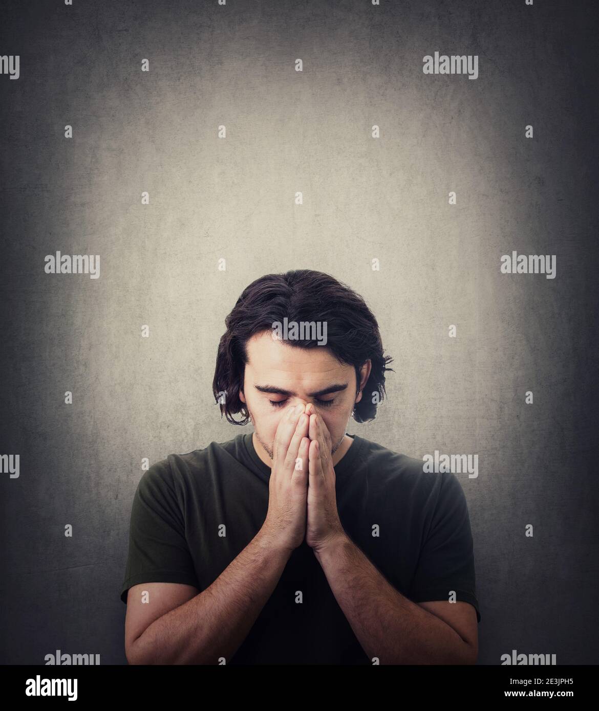Porträt von depressiven jungen Mann hält die Hände zusammen als Gebet, Augen geschlossen, Kopf nach unten isoliert auf grauem Hintergrund. Verzweifelter Kerl, der um Hilfe betet. Co Stockfoto