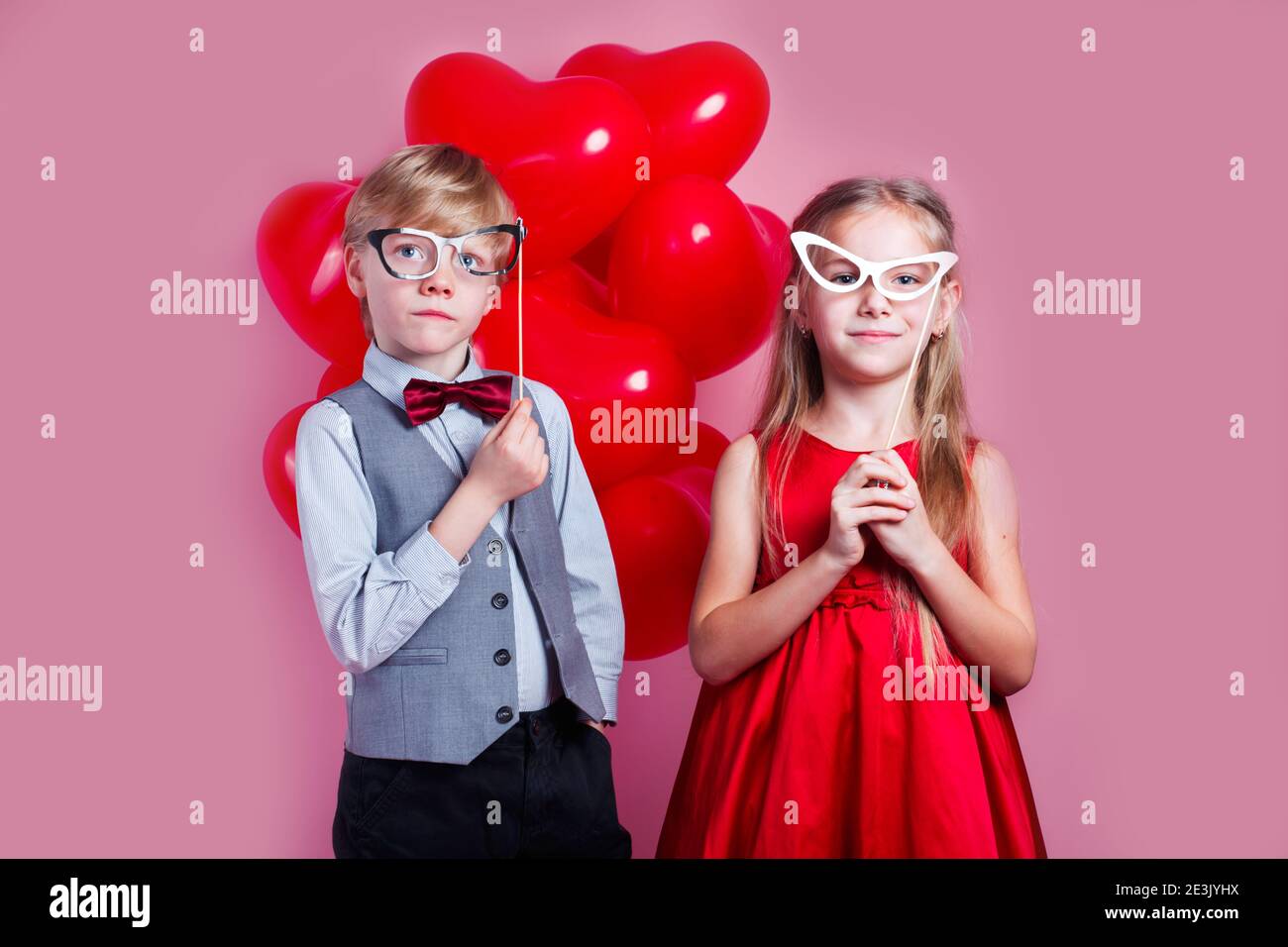 Valentinstag . Kleine liebevolle Paar Kinder in Pappgläsern mit roten Herz Ballons auf rosa Hintergrund. Stockfoto