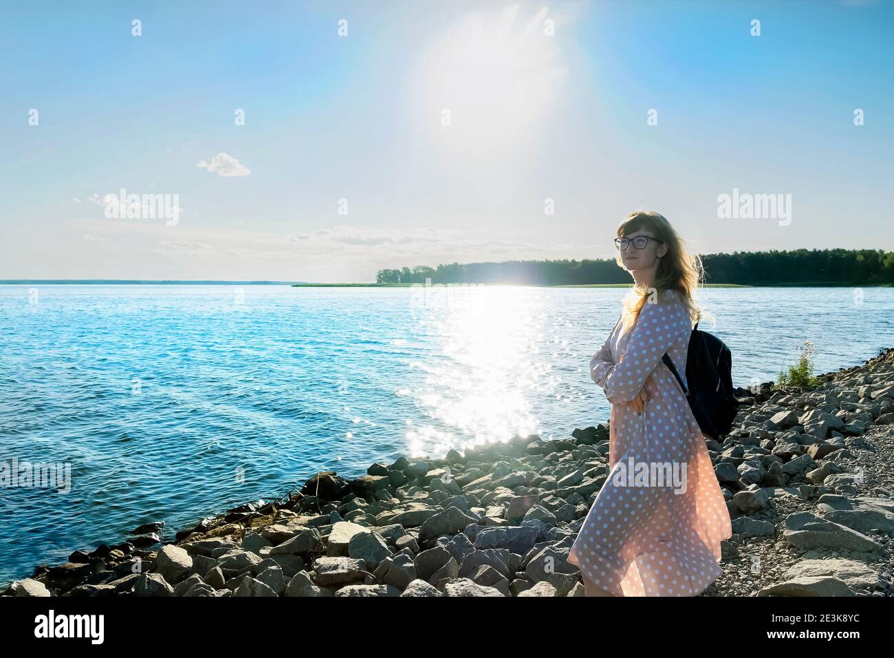 Frau in hellem Kleid steht auf einem Steinufer des Flusses oder See genießen das Sonnenlicht, Entspannung und Aussicht, geistige Gesundheit, weg von allem Stockfoto