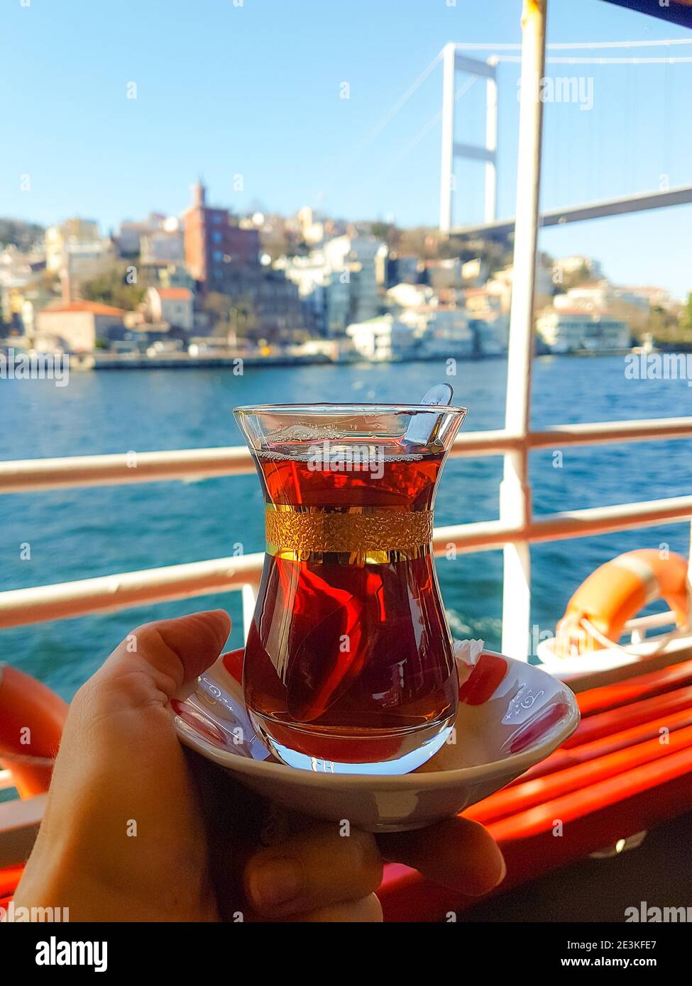 Traditioneller schwarzer türkischer Tee im Glas auf einem Teller in den Händen mit Blick auf die Bosporus-Meerenge und Istanbul-Hintergrund. Reise nach Türkei. Stockfoto