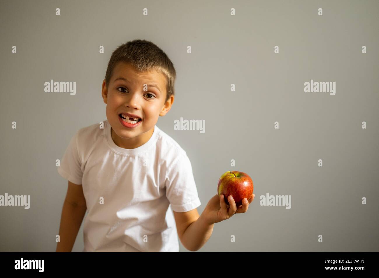 Junge ohne Milch Oberzahn in weißem T-Shirt hält einen Apfel in der Hand auf dem grauen Hintergrund. Stockfoto