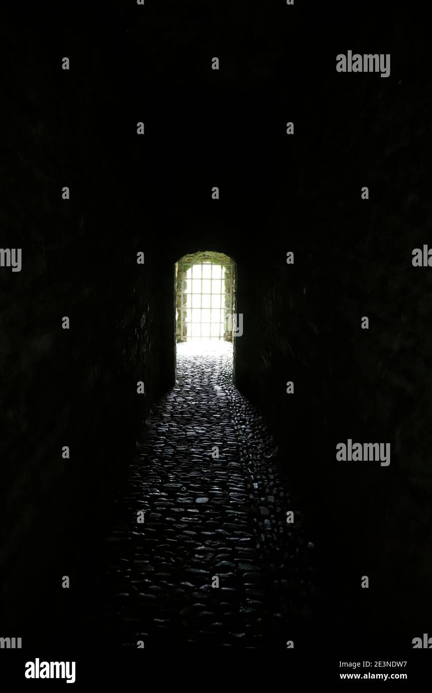 Dramatischer, dunkler, steingepflasterter Tunnel, der in einer hellen Öffnung endet, die durch Metallrost blockiert wird Stockfoto