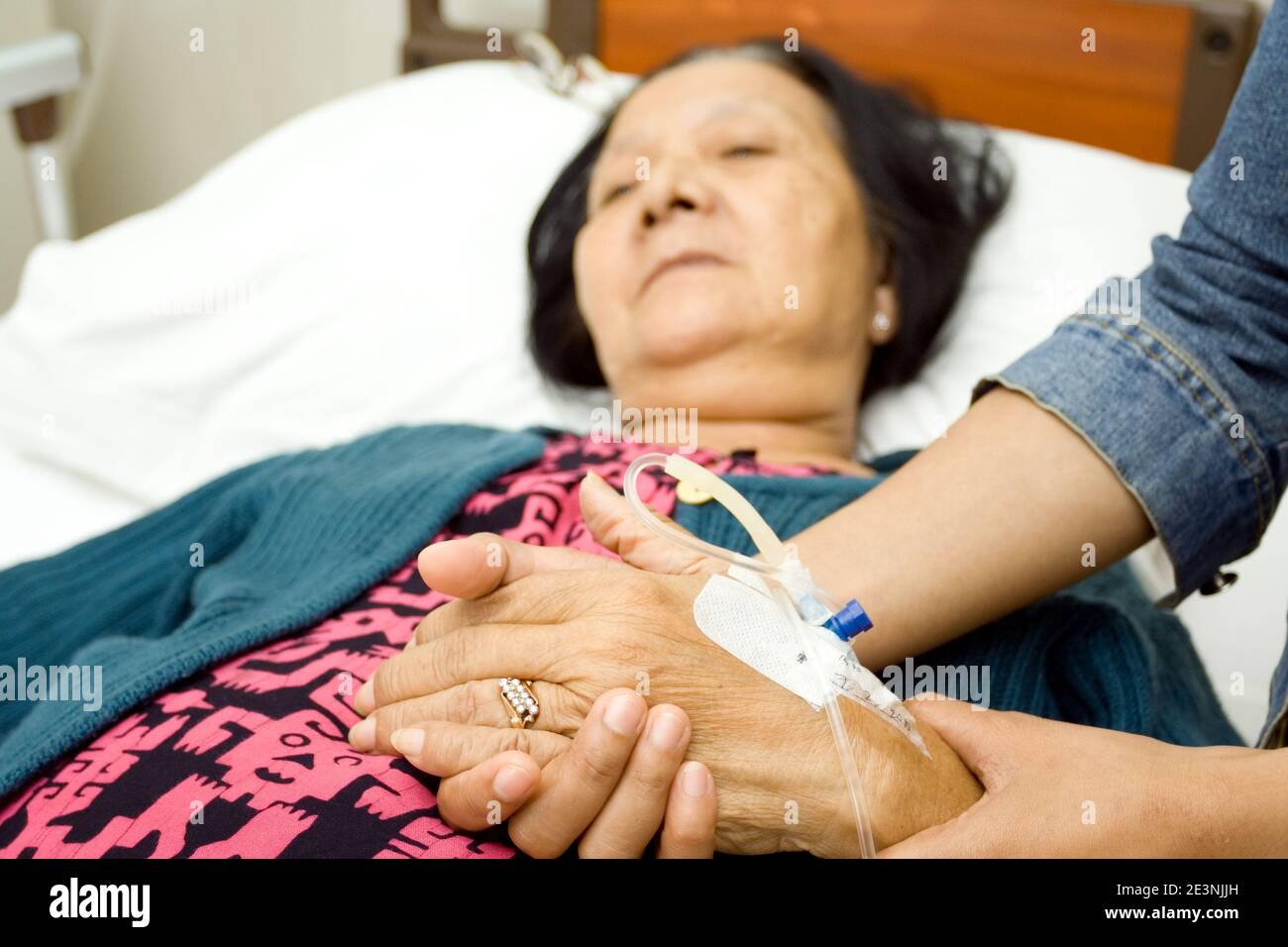 Eine ältere südostasiatische Frau, die wegen Krankheit ins Krankenhaus eingeliefert wurde, lag auf dem Krankenhausbett mit ihren Händen von ihrer Tochter gehalten Stockfoto