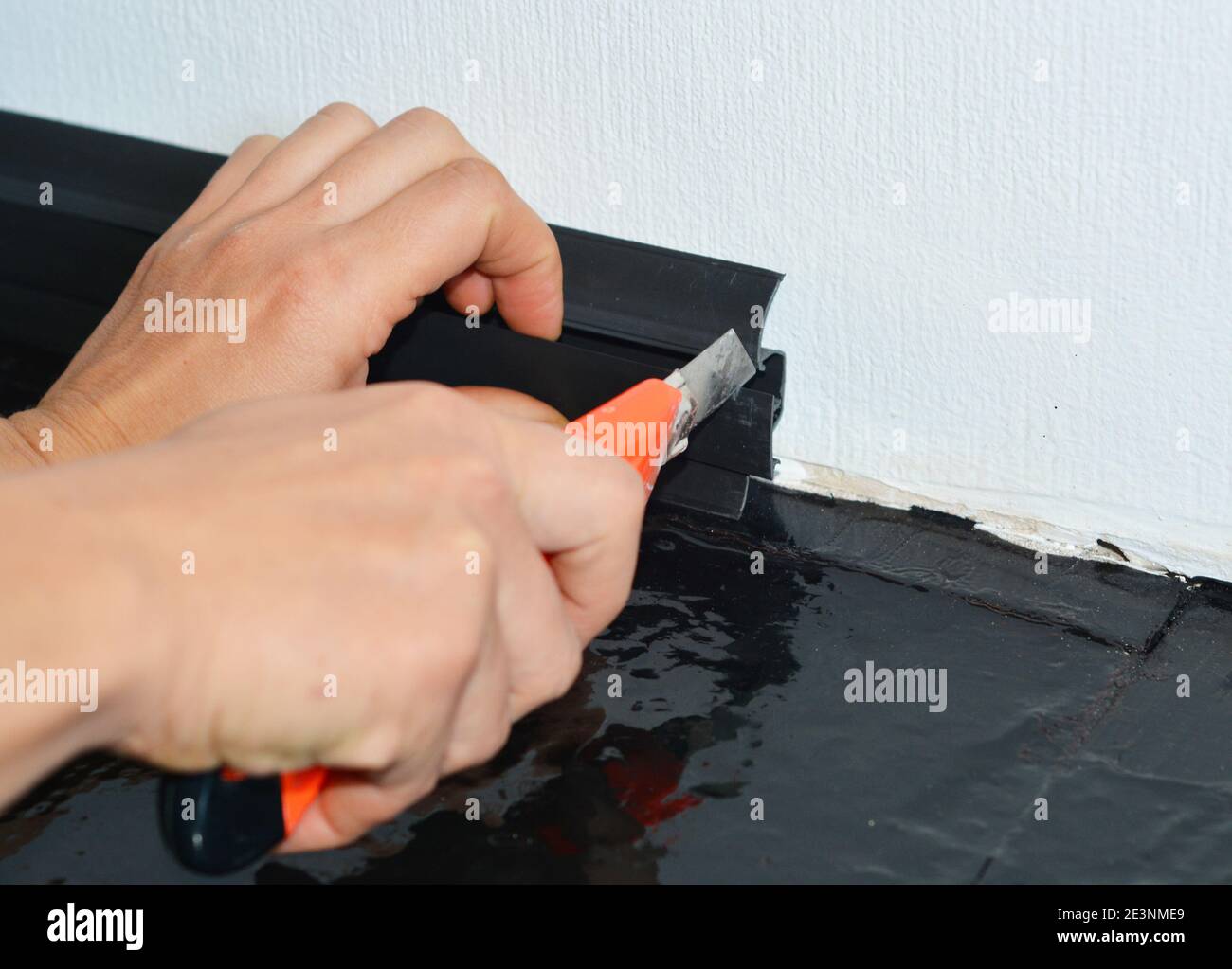Ein Handwerker installiert eine schwarze Kunststoff-Sockelleiste an einer weißen Wand und schneidet sie mit einem Gebrauchsmesser. Stockfoto