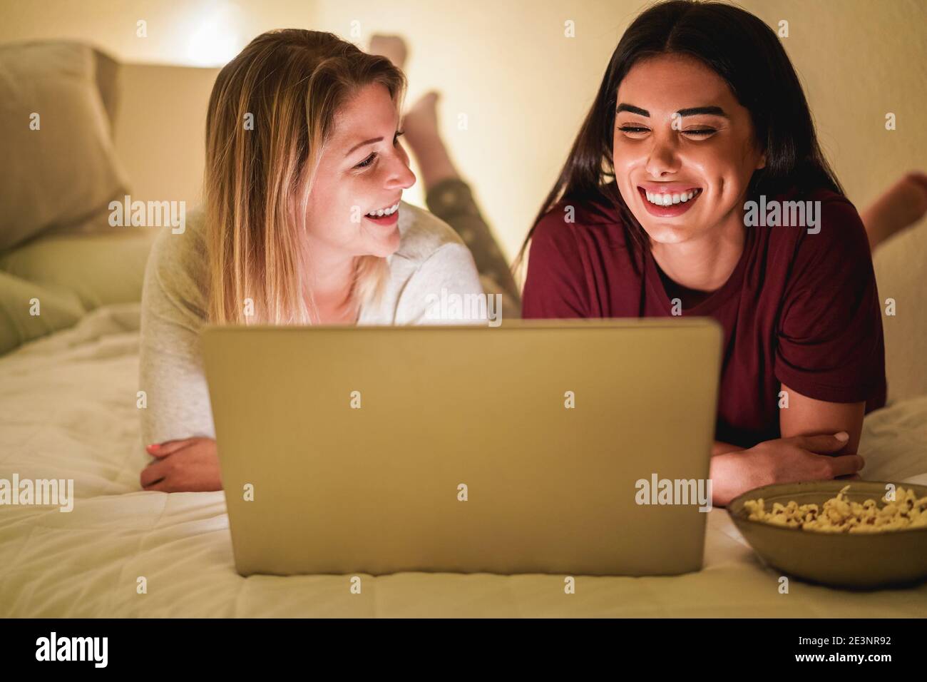 Zwei junge Freundinnen mit Laptop-Computer zusammen auf dem Bett Während Lockdown Isolation - Fokus auf das richtige Mädchen Stockfoto