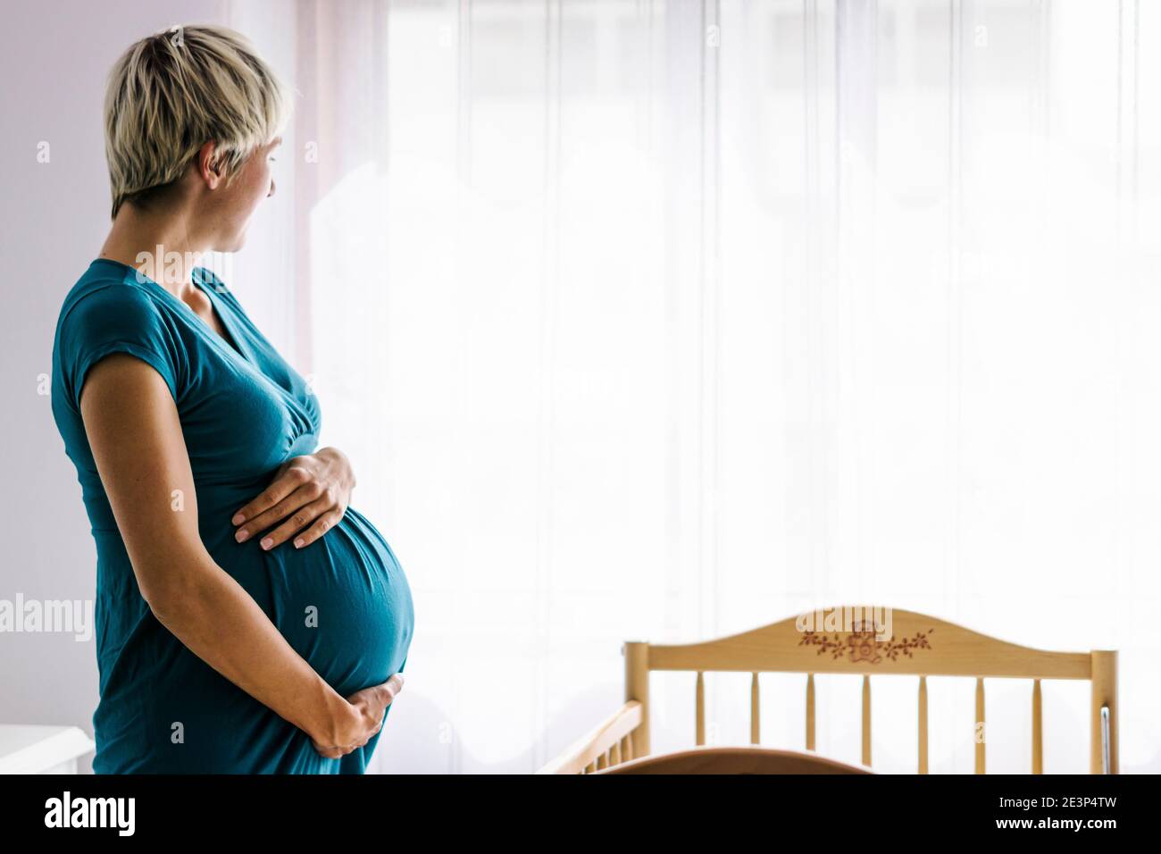 Schwangere Frau hält ihren Bauch von Kinderbett neben Das Fenster Stockfoto
