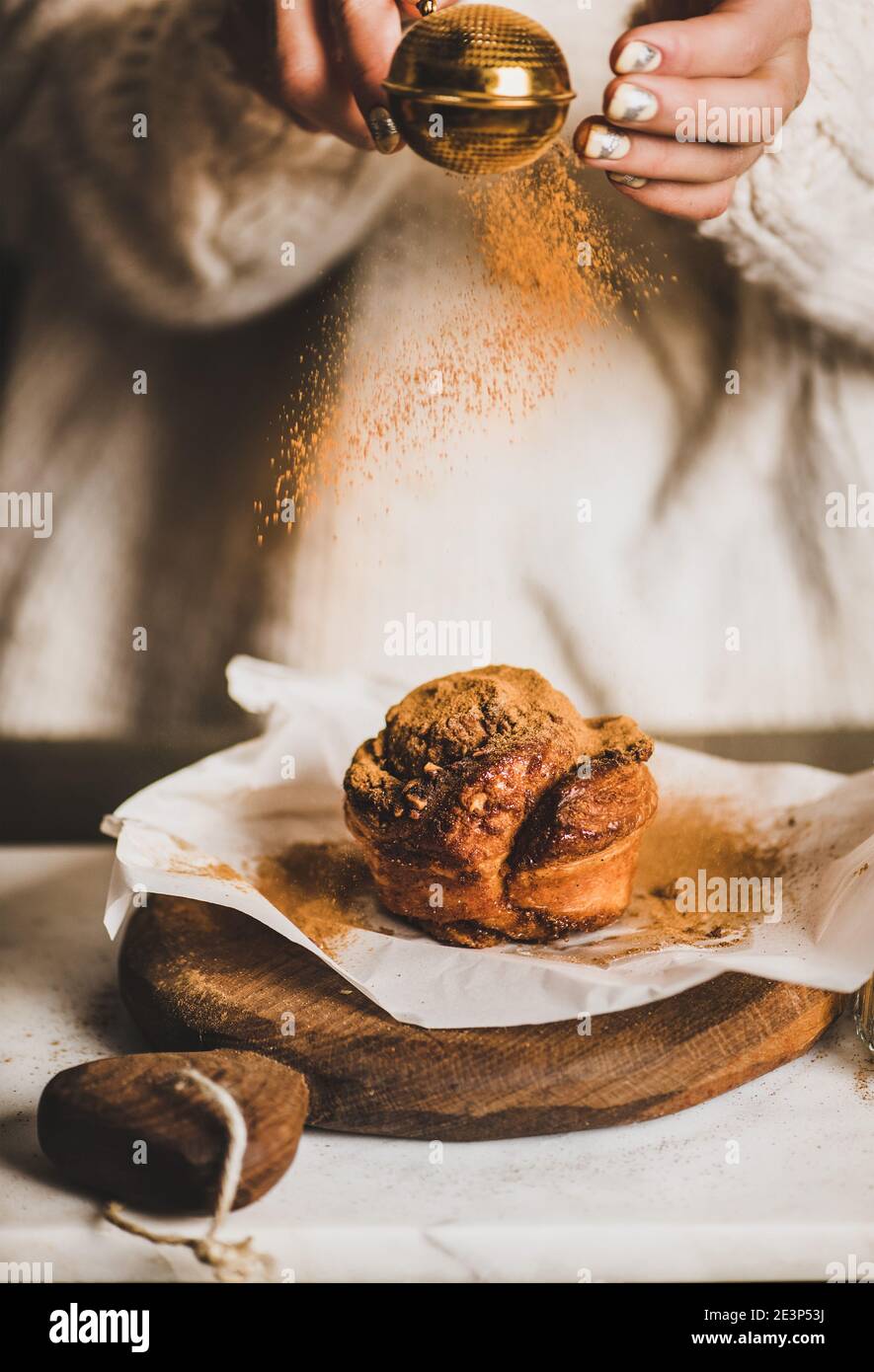 Weibliche Hände bestreuen gemahlenes Zimtpulver über süßes warmes Zimt-Karamell-Brötchen auf rustikalem Holzbrett, selektiver Fokus. Winter Dessert, Komfort-Food-Konzept Stockfoto