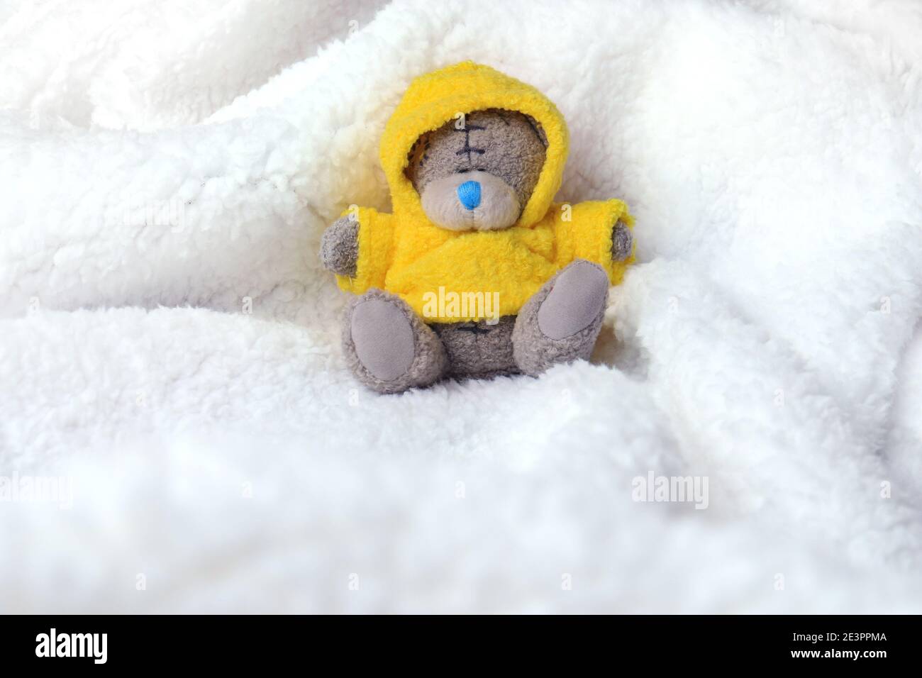 Ein kleiner Teddybär mit gelbem Mantel, der in einer weißen Felldecke sitzt. Stockfoto