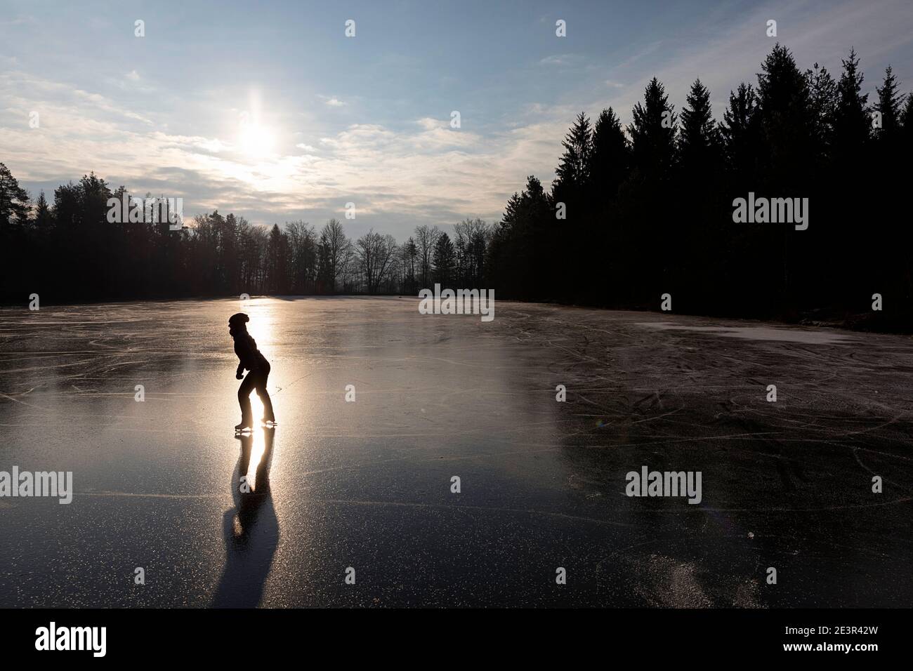 Silhouette einer Frau Eislaufen auf gefrorenem See, beleuchtet von der Sonne von hinten bei Sonnenaufgang, Slowenien Stockfoto