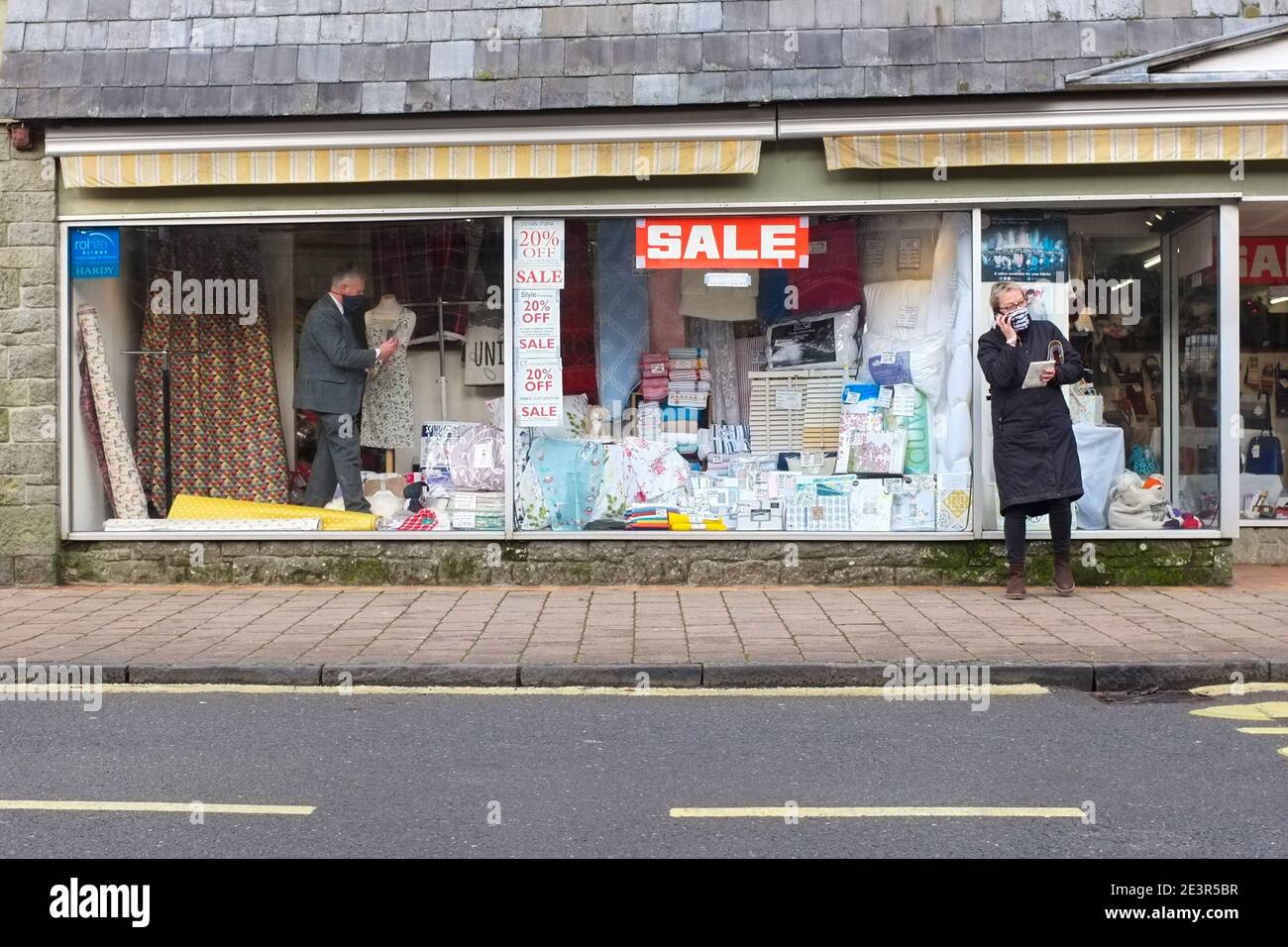 Großbritannien gewöhnt sich an die Einschränkungen des Coronavirus. Ein maskierter Shopper außerhalb eines Shops, in dem ein maskierter Assistent die Anzeige des Verkaufsfensters vorbereitet. Dezember 2020. Stockfoto