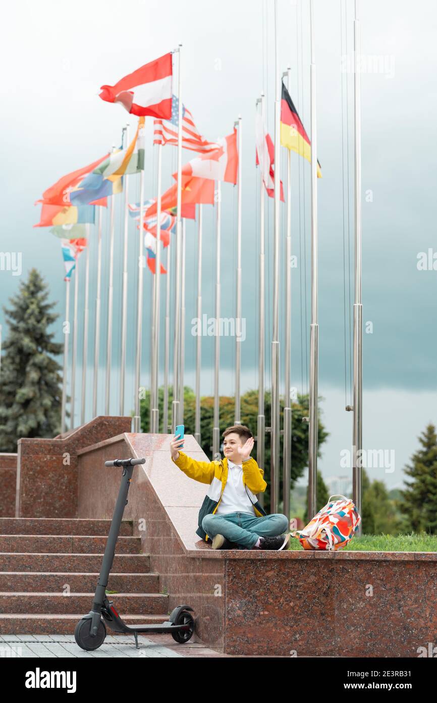 Teenager sitzt in der Nähe des Verwaltungsgebäudes vor dem Hintergrund von Landesflaggen und Wellen in das Smartphone. Elektroroller Fahrer Konzept Stockfoto