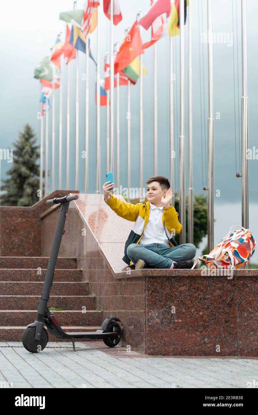 Teenager sitzt in der Nähe des Verwaltungsgebäudes vor dem Hintergrund von Landesflaggen und Wellen in das Smartphone. Elektroroller Fahrer Konzept Stockfoto