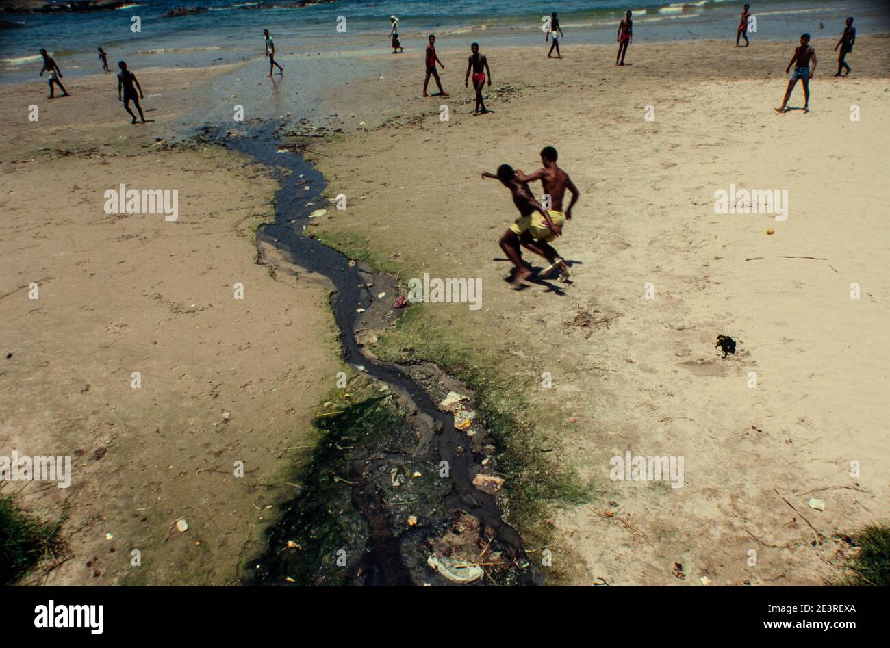 Schwarze junge Männer spielen Fußball neben offenen Abwässern, die direkt am Strand von Itapoa, Salvador City, Bahia State, im Nordosten Brasiliens entsorgt werden. Stockfoto