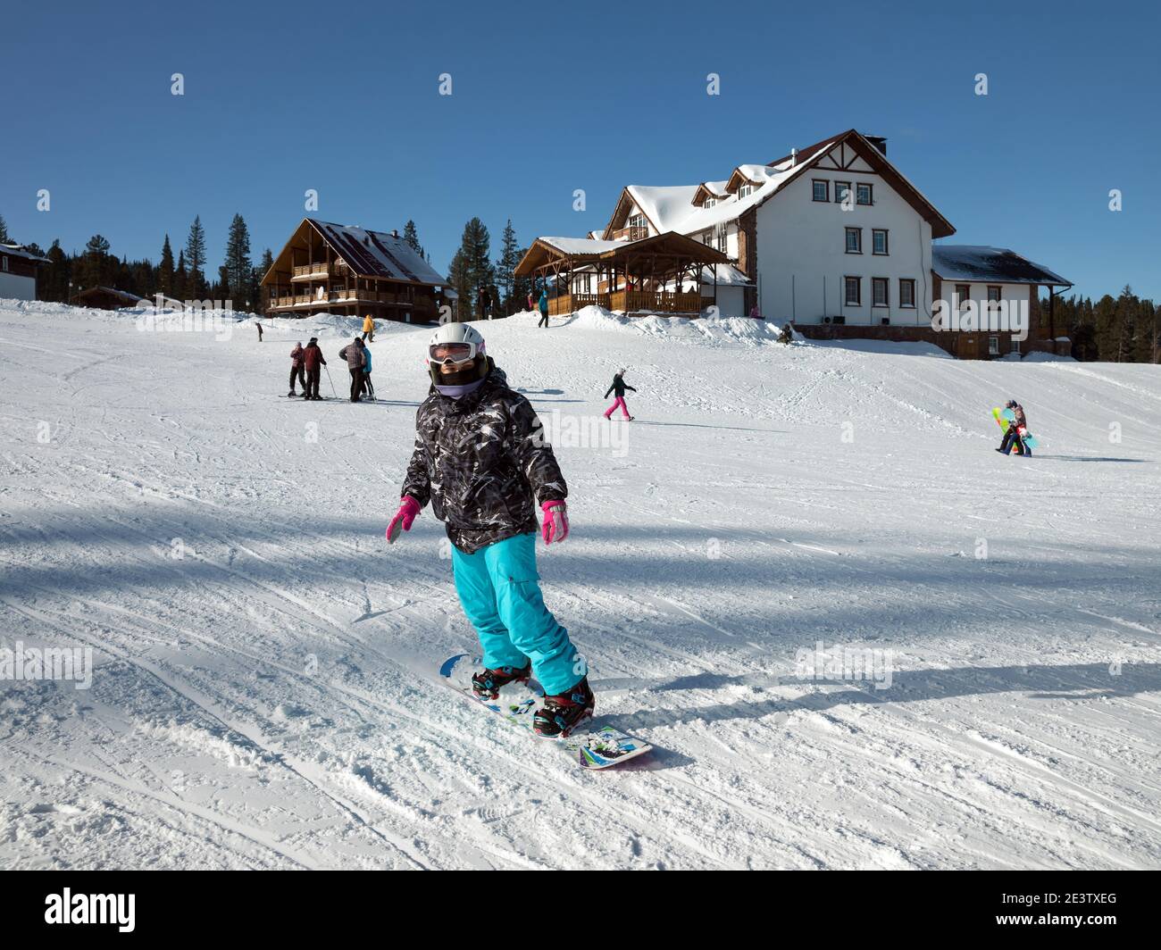 Eine junge Frau geht an einem sonnigen Wintertag mit dem Snowboard vor der Kulisse der Gebäude des Skigebiets den Berg hinunter. Stockfoto