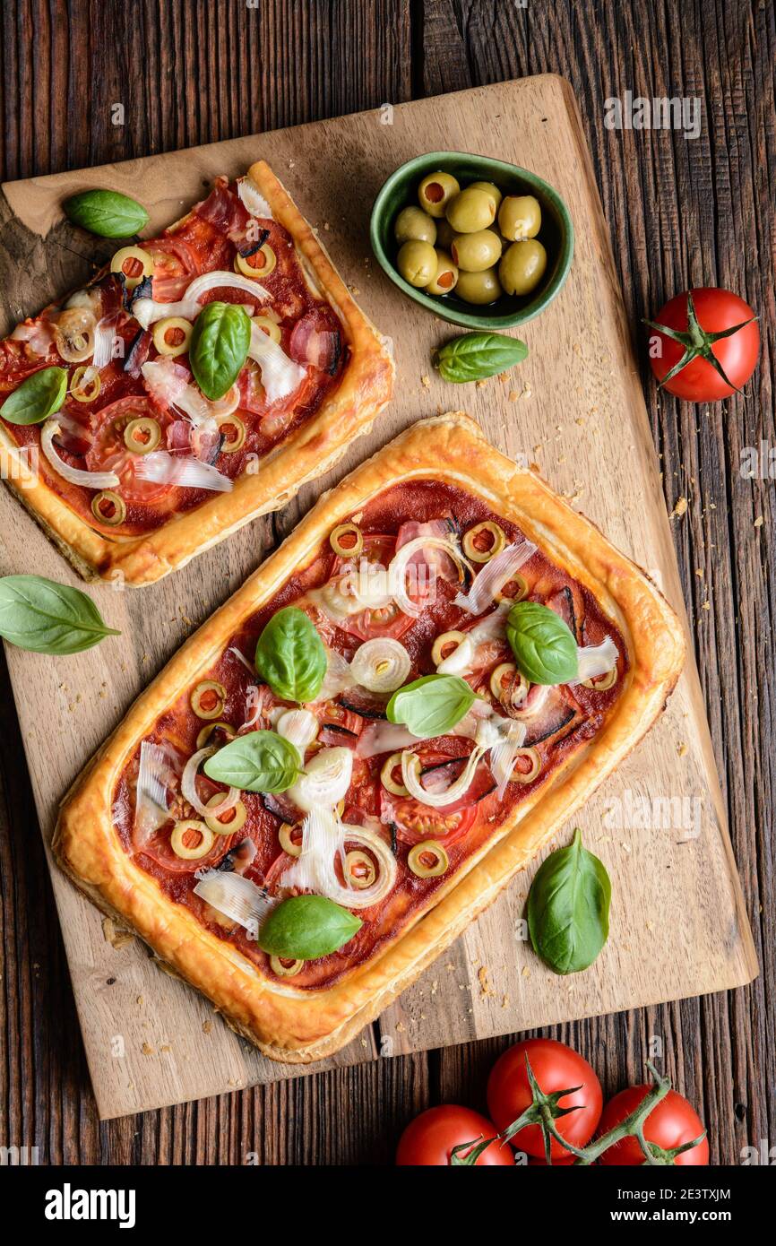 Hausgemachte Blätterteig-Pizza mit Speckscheiben, Tomaten, grünen Oliven, Käse und Zwiebeln, gekrönt mit Basilikumblättern auf rustikalem Holzhintergrund Stockfoto