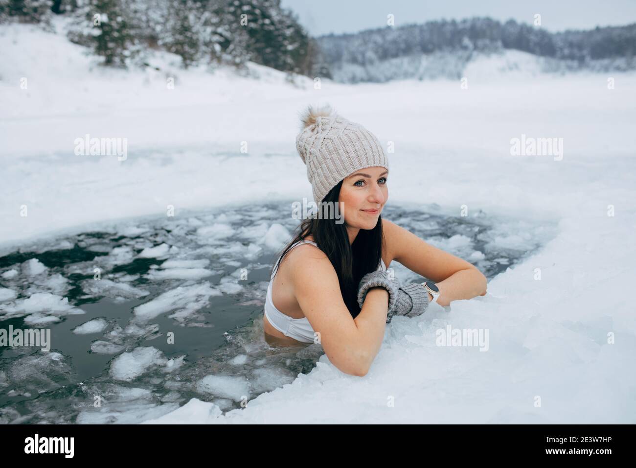 Schwimmen im Winter. Frau in gefrorenem See Eisloch. Wie man im kalten Wasser schwimmt. Schöne junge Frau lächelt. Badekleidung mit Mütze und Handschuhen. Stockfoto