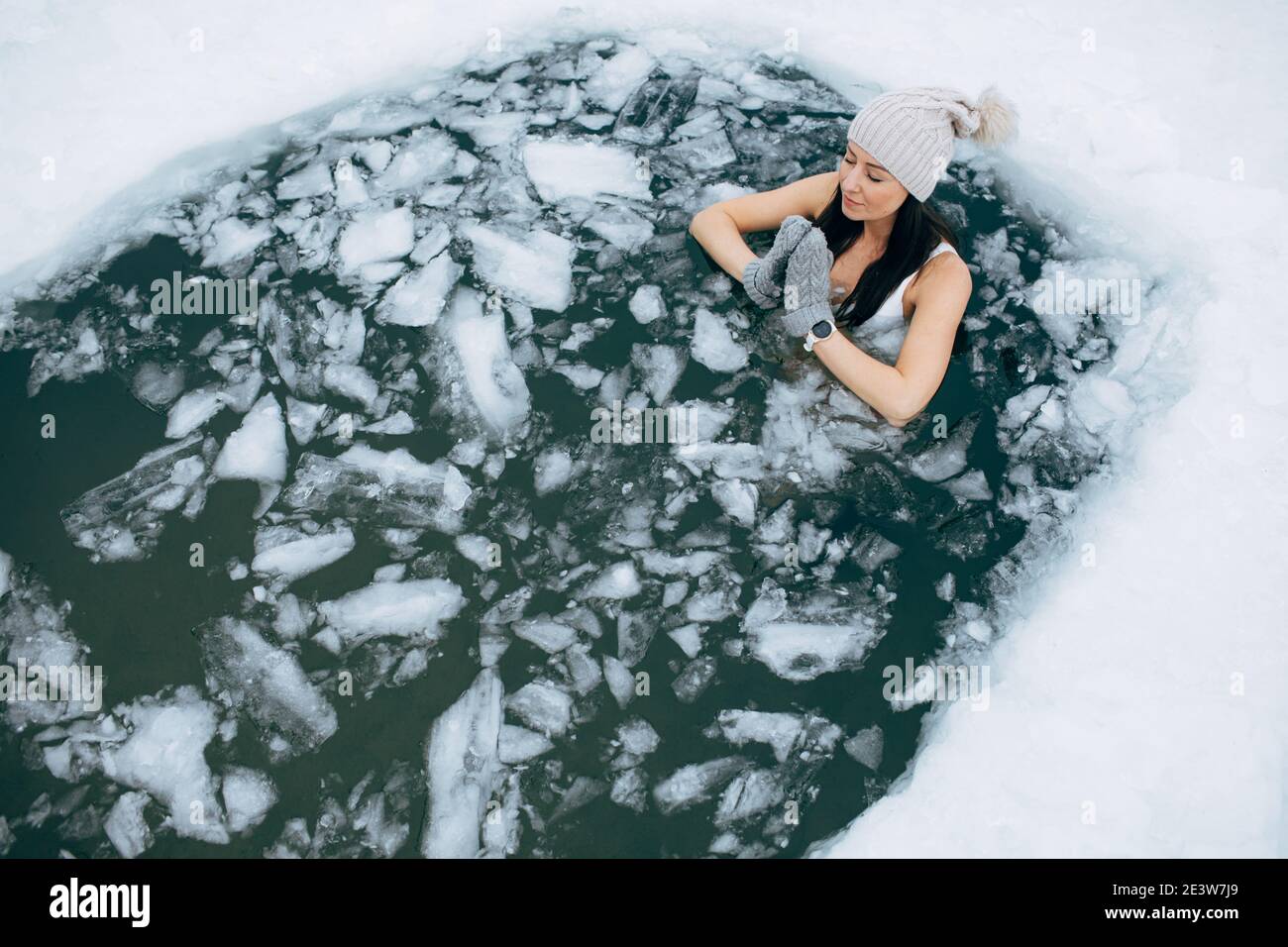 Schwimmen im Winter. Frau in gefrorenem See Eisloch. Wie man im kalten Wasser schwimmt. Schöne junge Frau in Zen-Meditation. Badekleidung mit Mütze und Handschuhen. Stockfoto