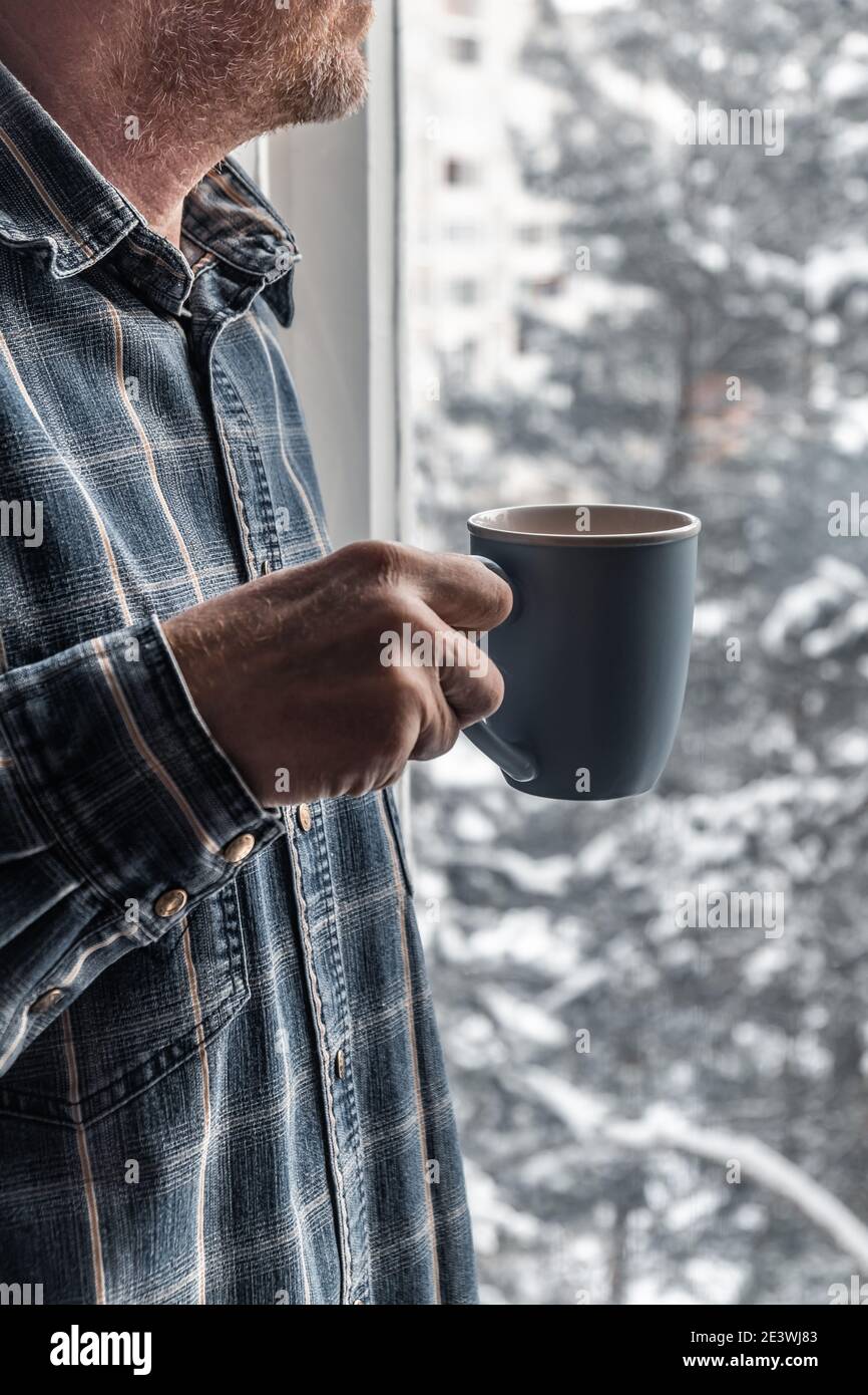 Ein blauer Becher mit Kaffee in den Händen eines Mannes vor dem Hintergrund eines Fensters mit schneebedeckten Bäumen an einem Wintermorgen. Wärmende Getränk im Winter. Stockfoto